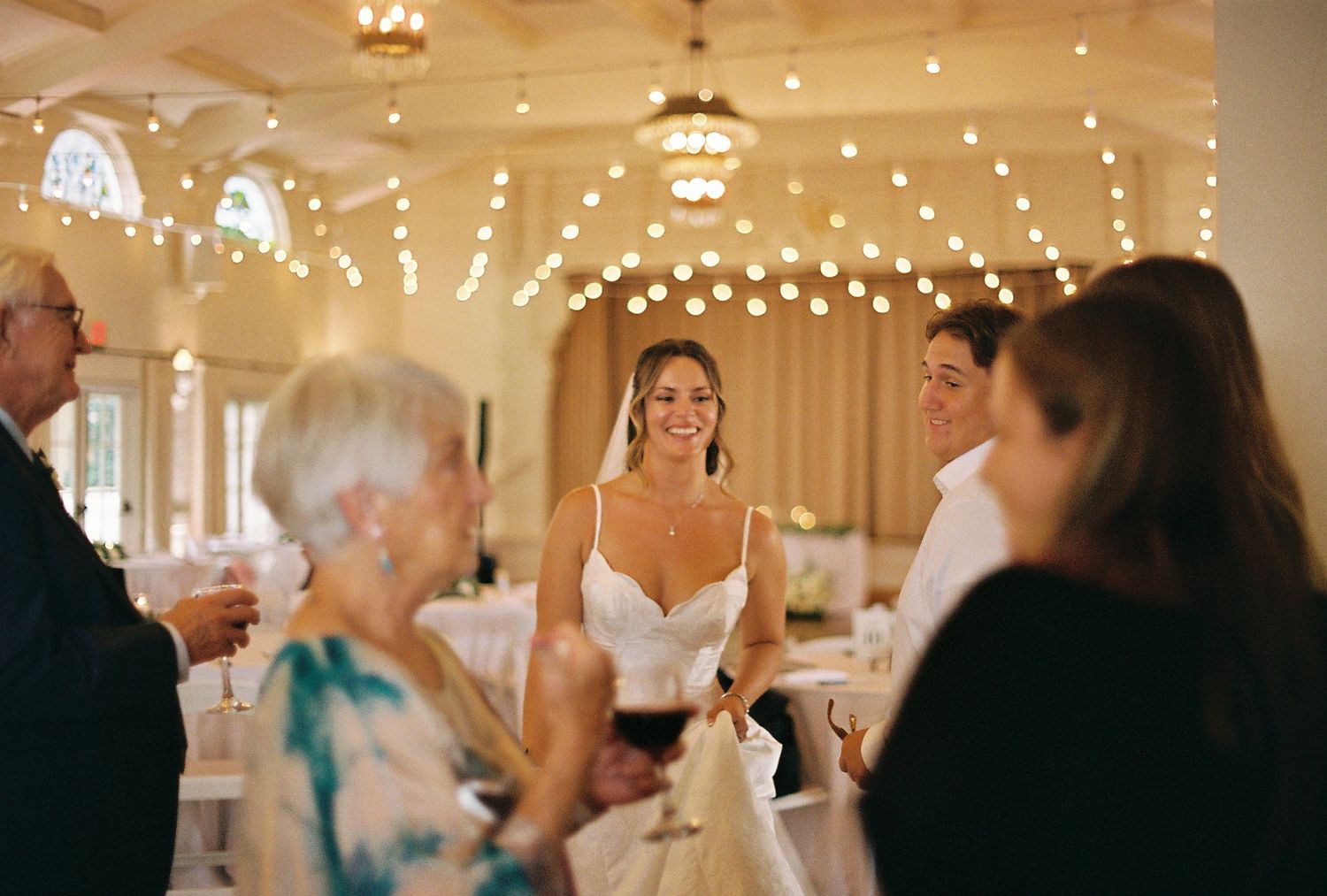 Bride and groom smiling with guests at elegant wedding reception venue with string lights.