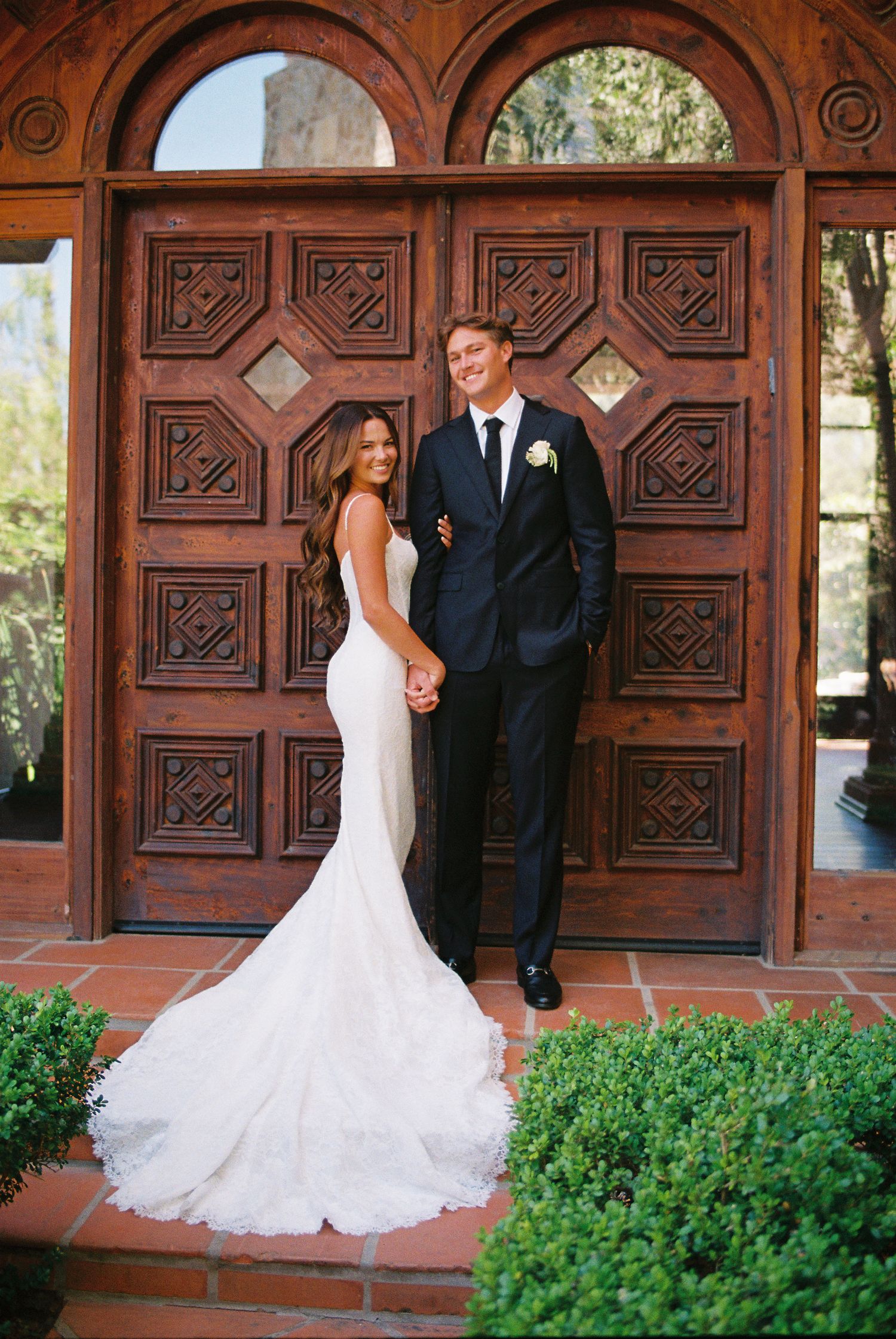Bride in mermaid gown and groom in black tuxedo pose before ornate carved wooden doors at elegant wedding venue.