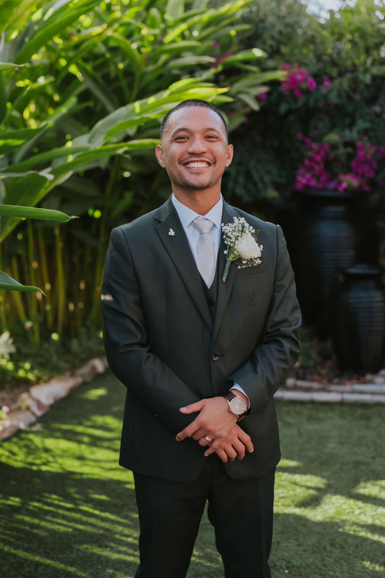 A groom in a dark suit poses in a lush tropical garden setting with bright flowers and greenery.