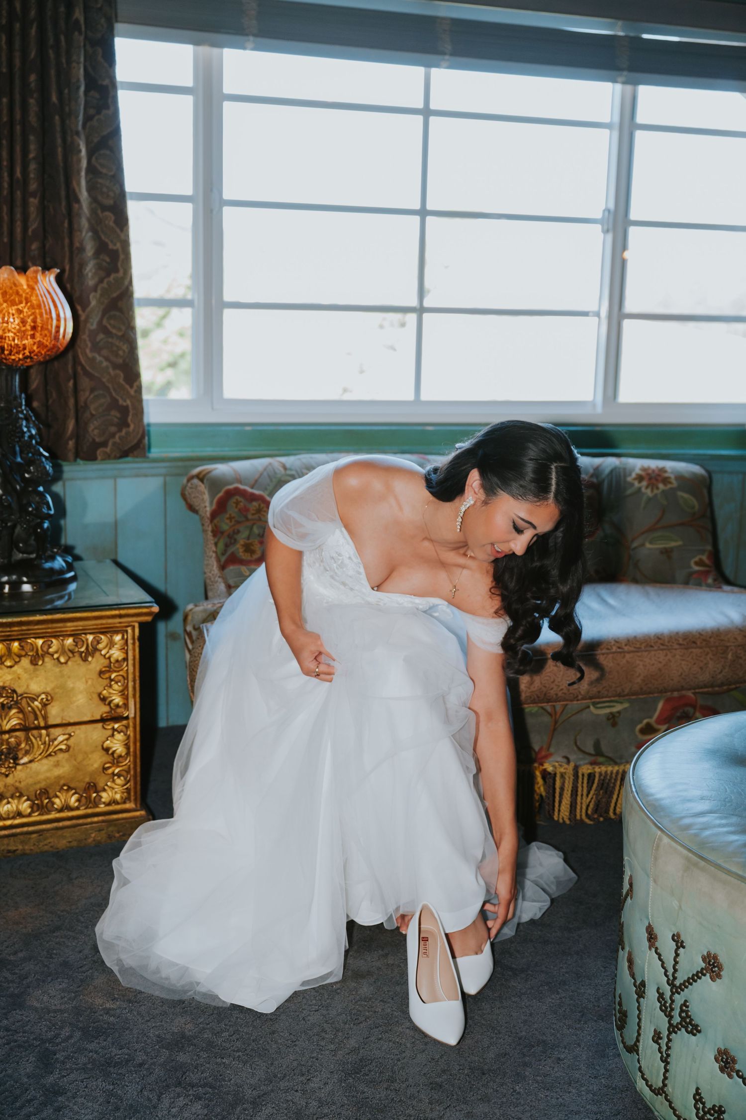 Bride in white wedding dress adjusts her shoe while sitting indoors near ornate furniture.