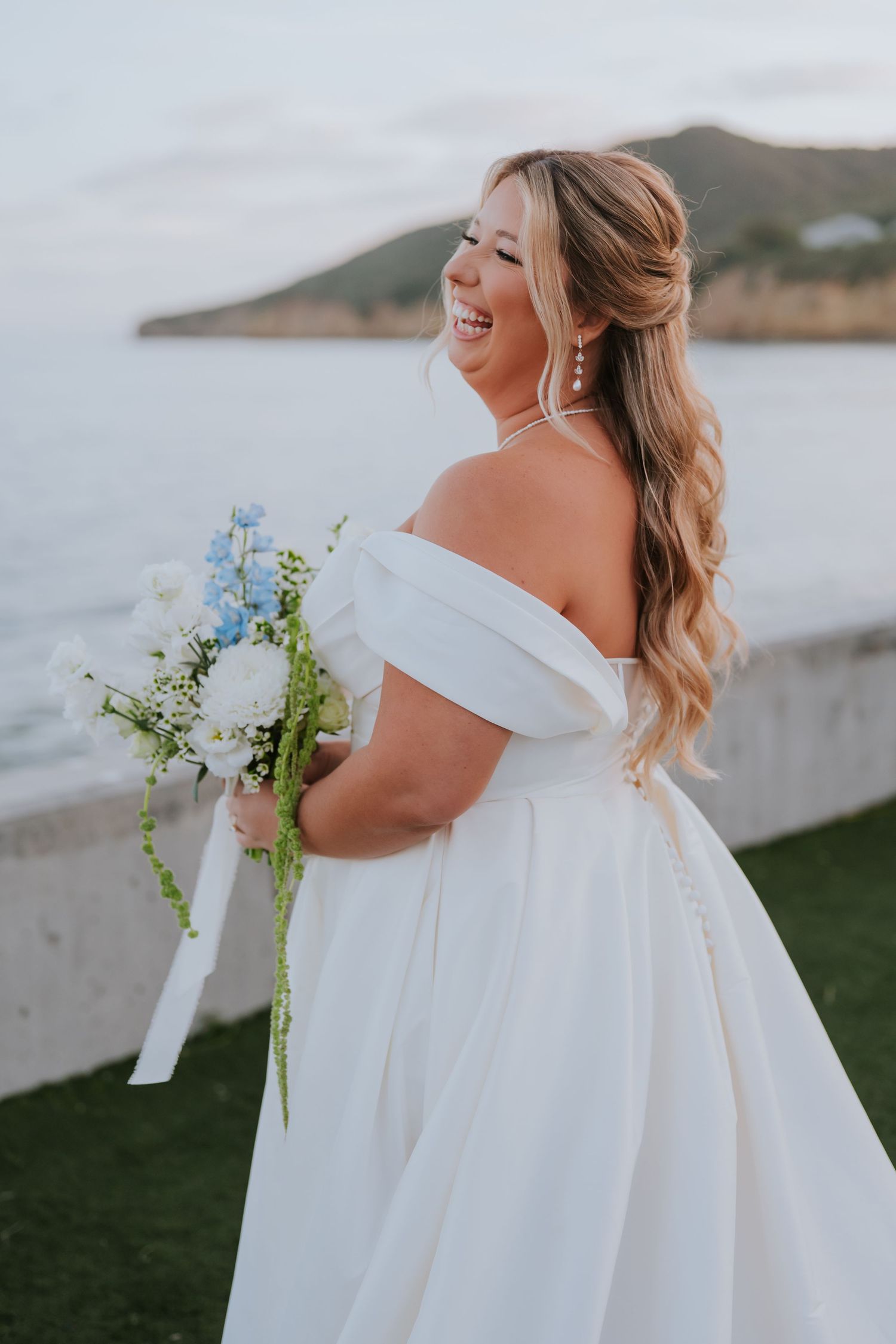 Smiling bride in off-shoulder white gown holds blue and white bouquet, standing by the sea with hills in background.