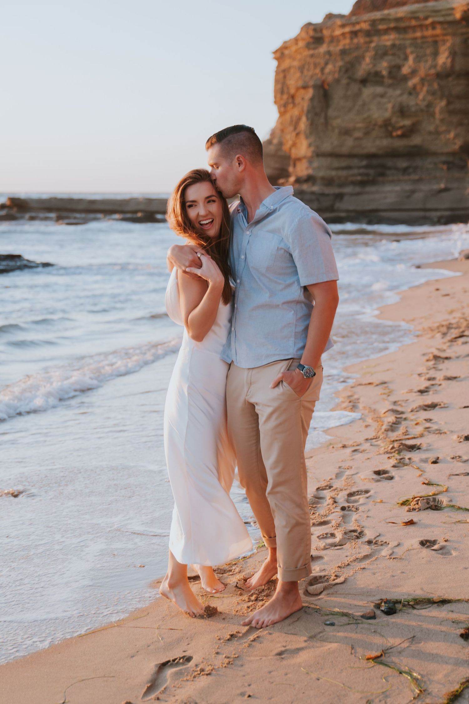 Couple embracing on a sandy beach at sunset, woman in white dress smiling, man kissing her cheek near rocky cliffs.