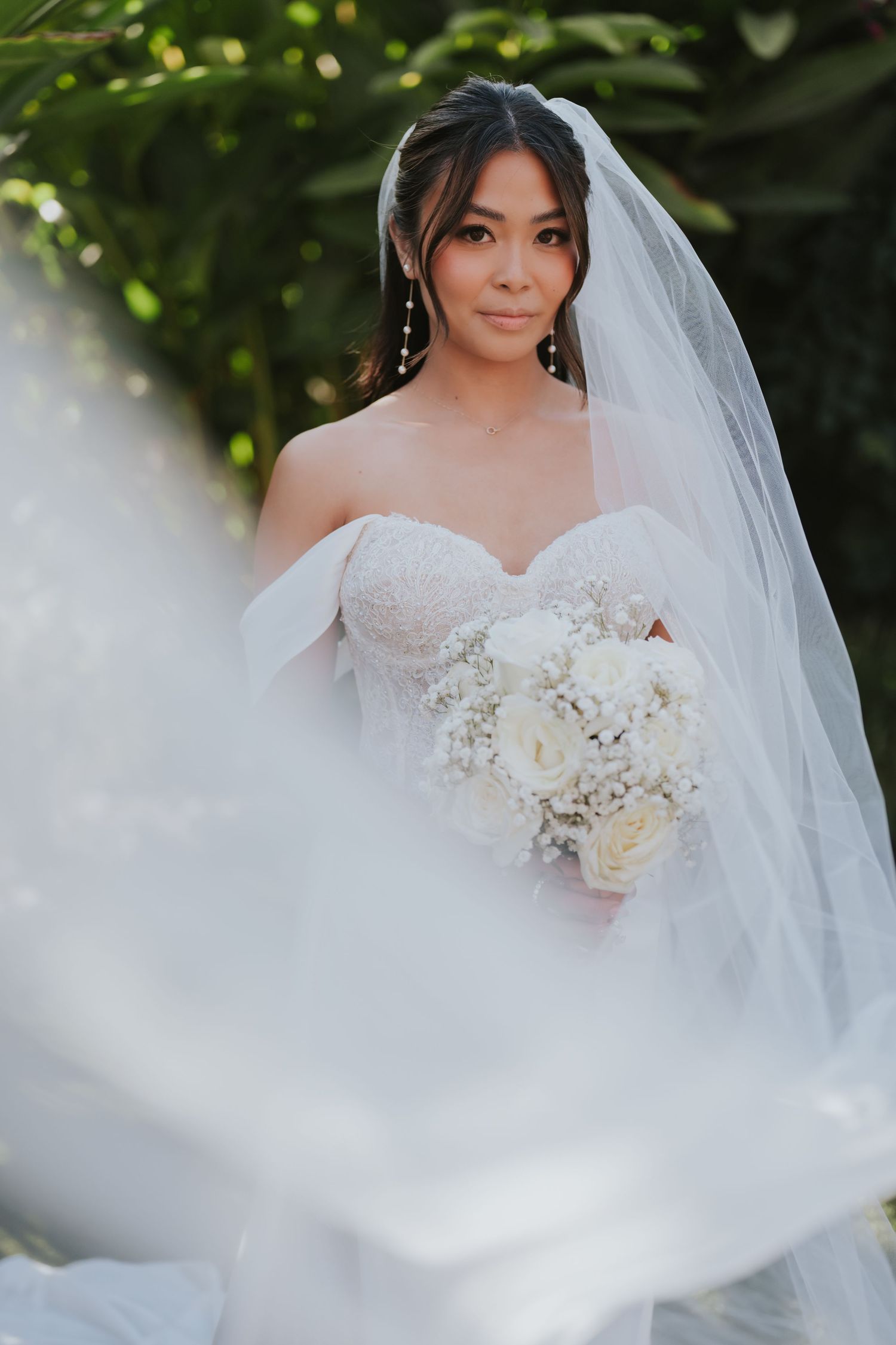 A bride in a strapless white wedding gown holds an elegant white rose bouquet while wearing a flowing veil.