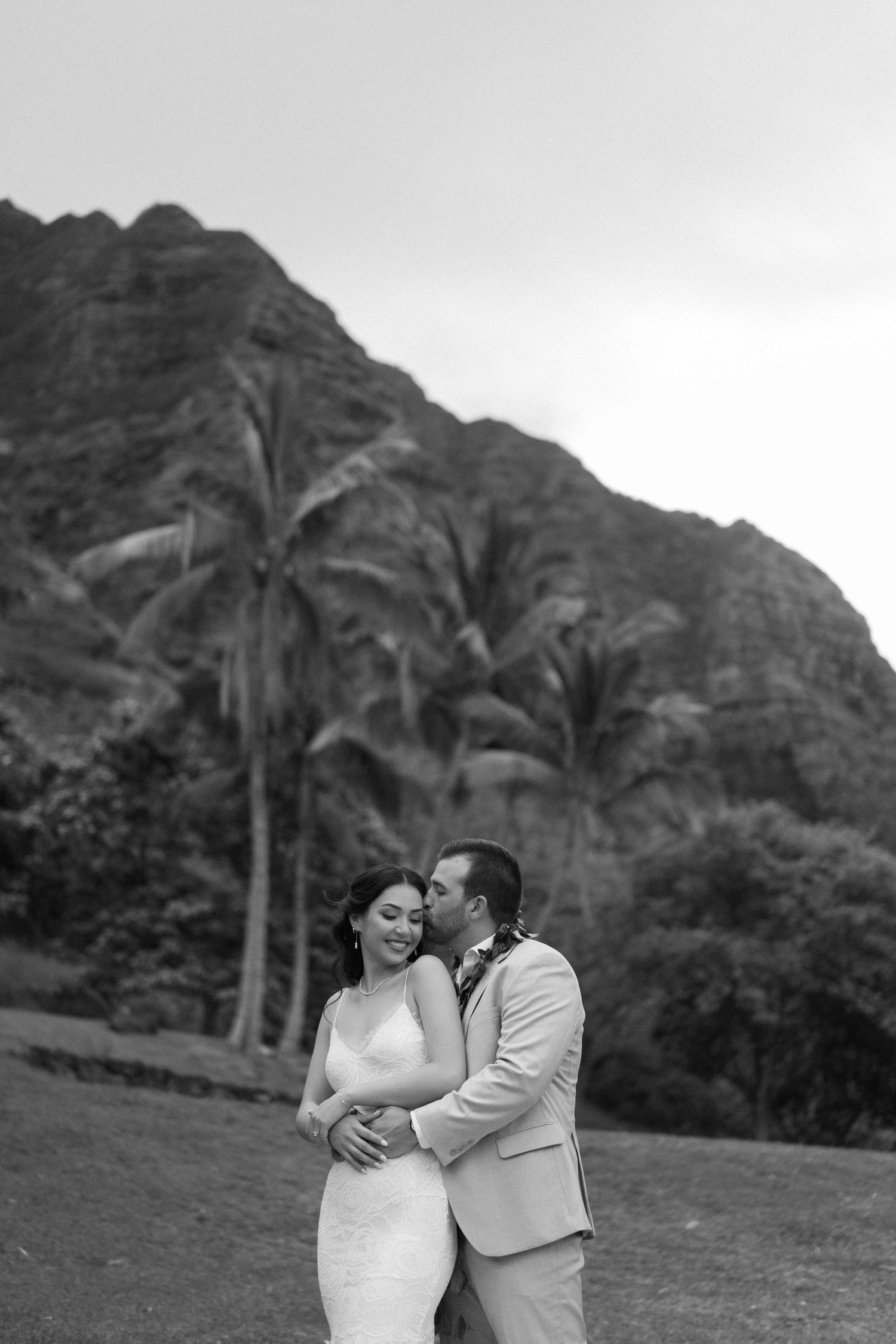 Black and white portrait of newlyweds embracing with tropical mountain backdrop.