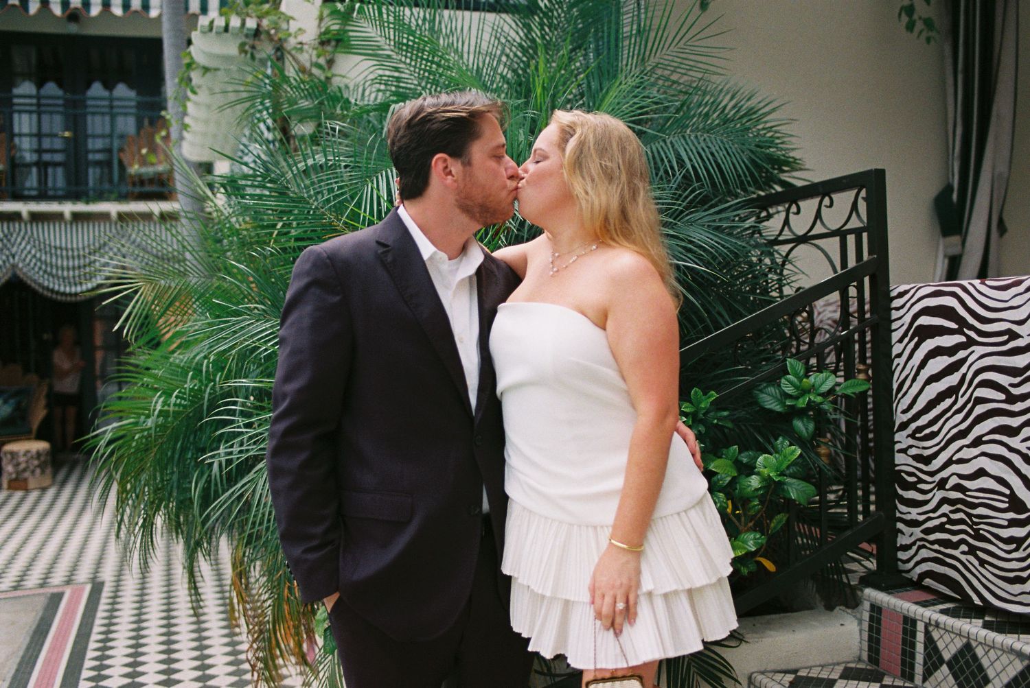 Couple sharing a romantic kiss outdoors, woman in white strapless dress and man in dark suit near tropical plants.