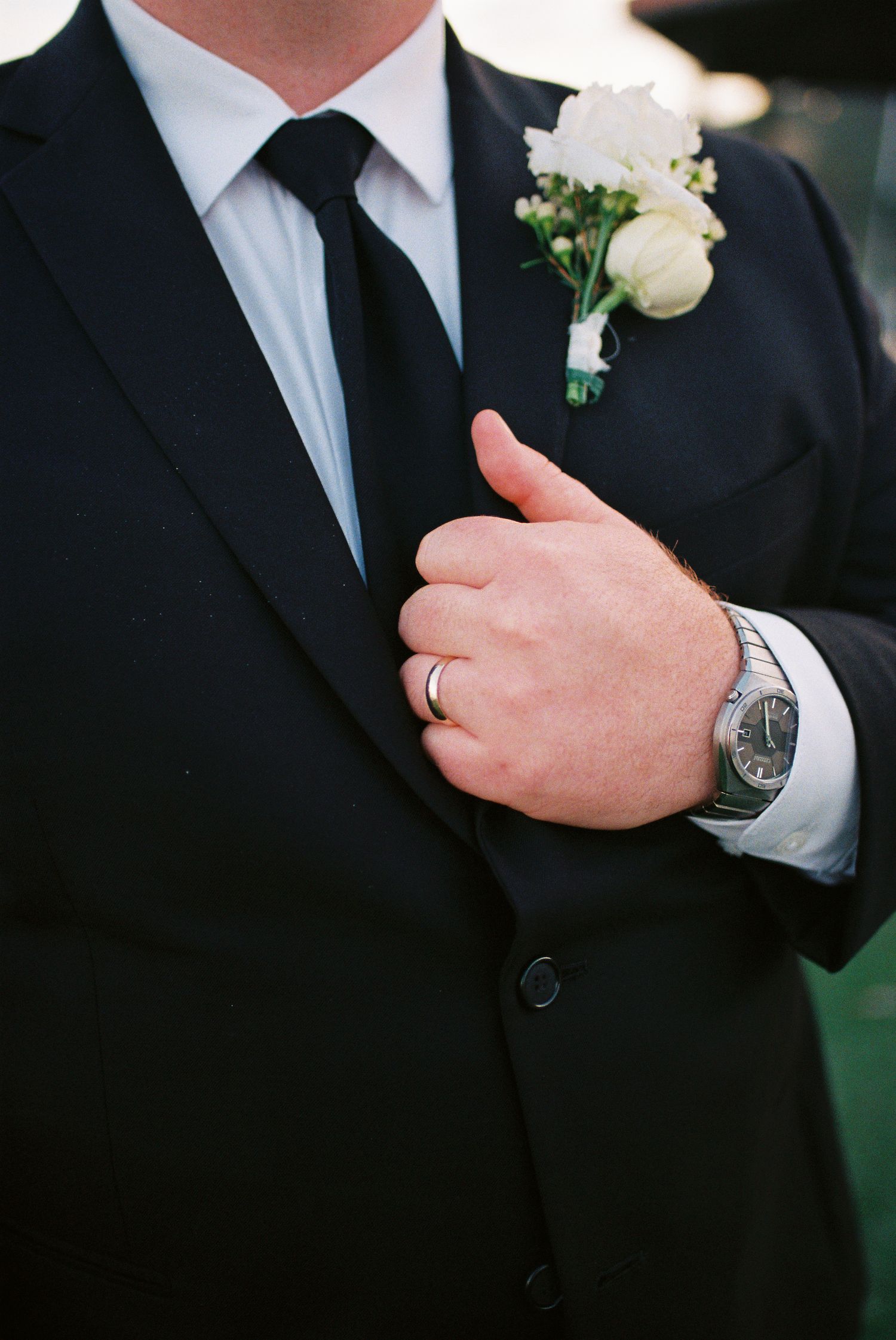 Groom in black suit with white rose boutonniere, silver watch, and wedding ring giving thumbs up gesture.