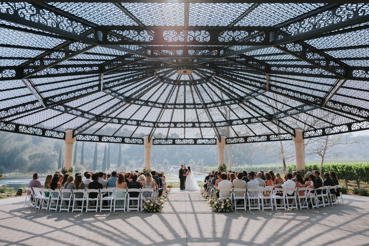 Elegant outdoor wedding ceremony under ornate glass dome pavilion with white chairs and guests gathered around the altar.