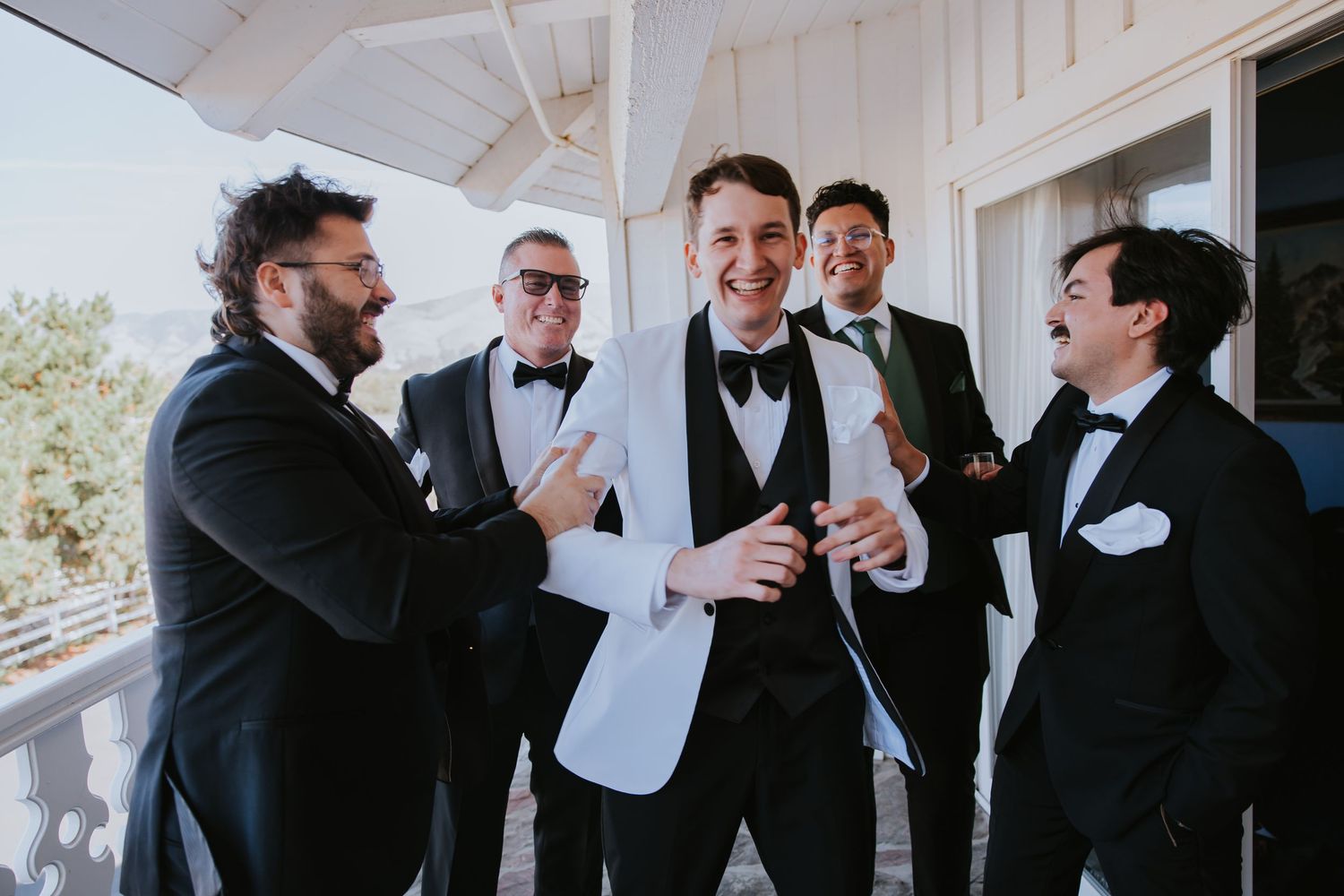 Wedding party members in black tuxedos share candid moments while getting ready indoors.