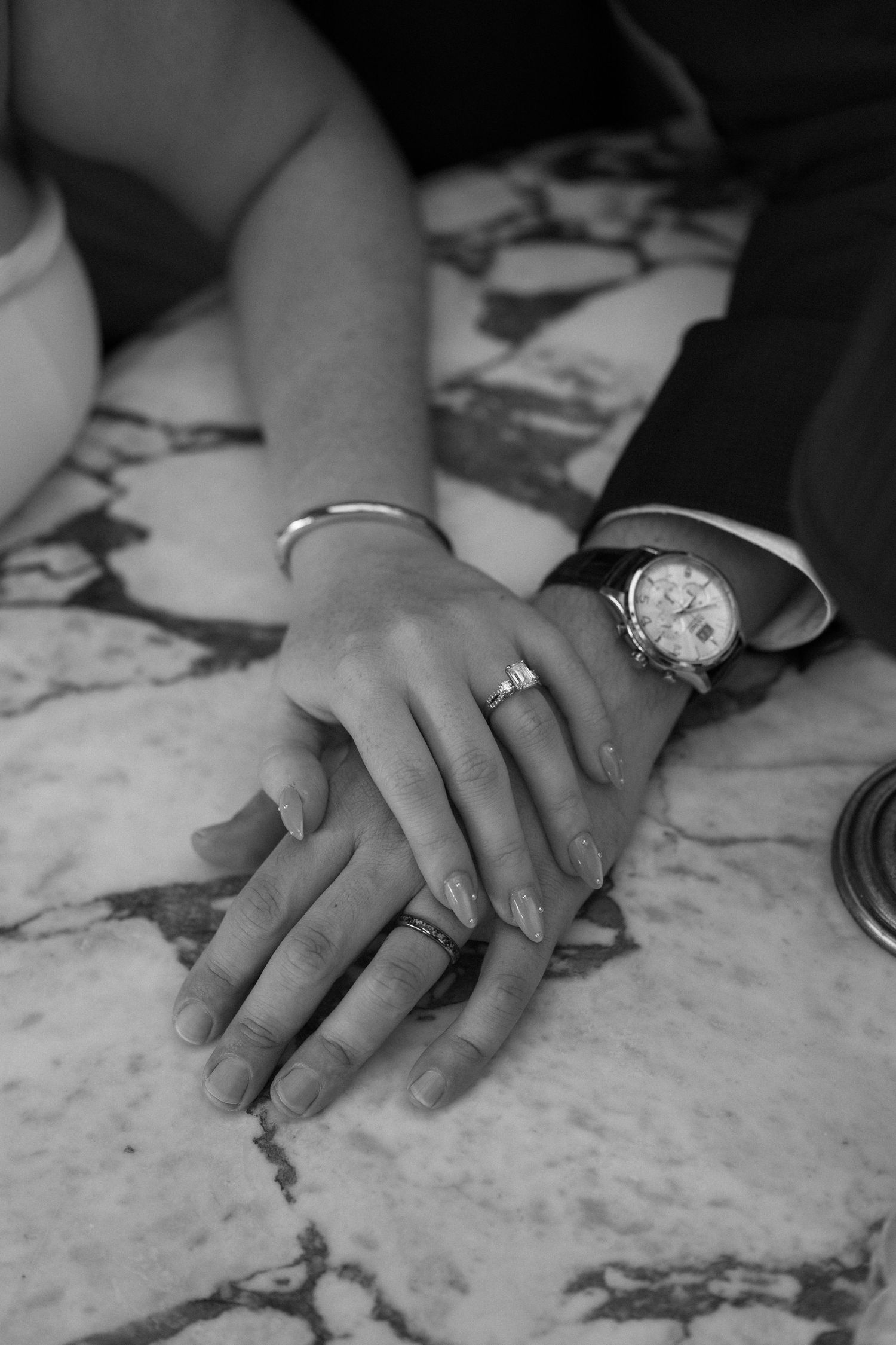 Close-up black and white photo of couple's hands with engagement ring and watch on marble surface.