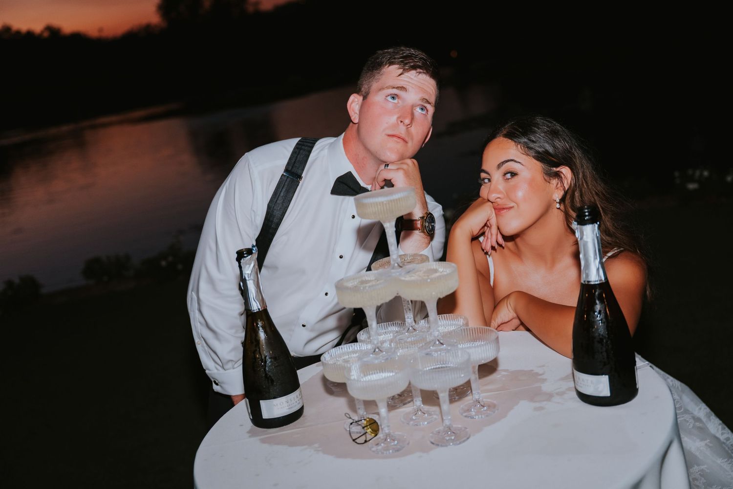 Bride and groom posing beside a champagne tower at sunset, with bottles of champagne on a white table outdoors.