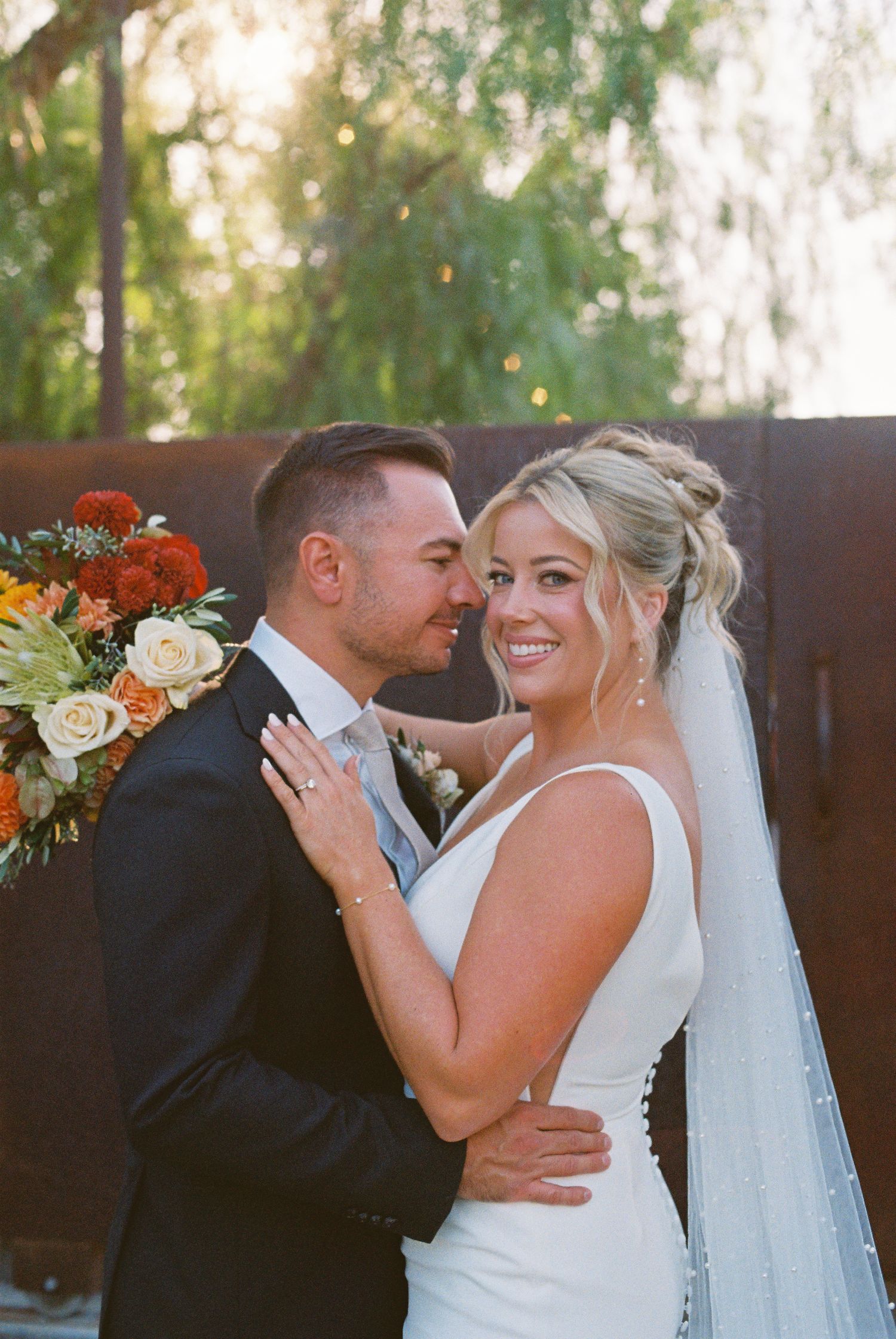 Smiling bride in white gown and veil embraces groom in black suit, with autumn floral bouquet outdoors.