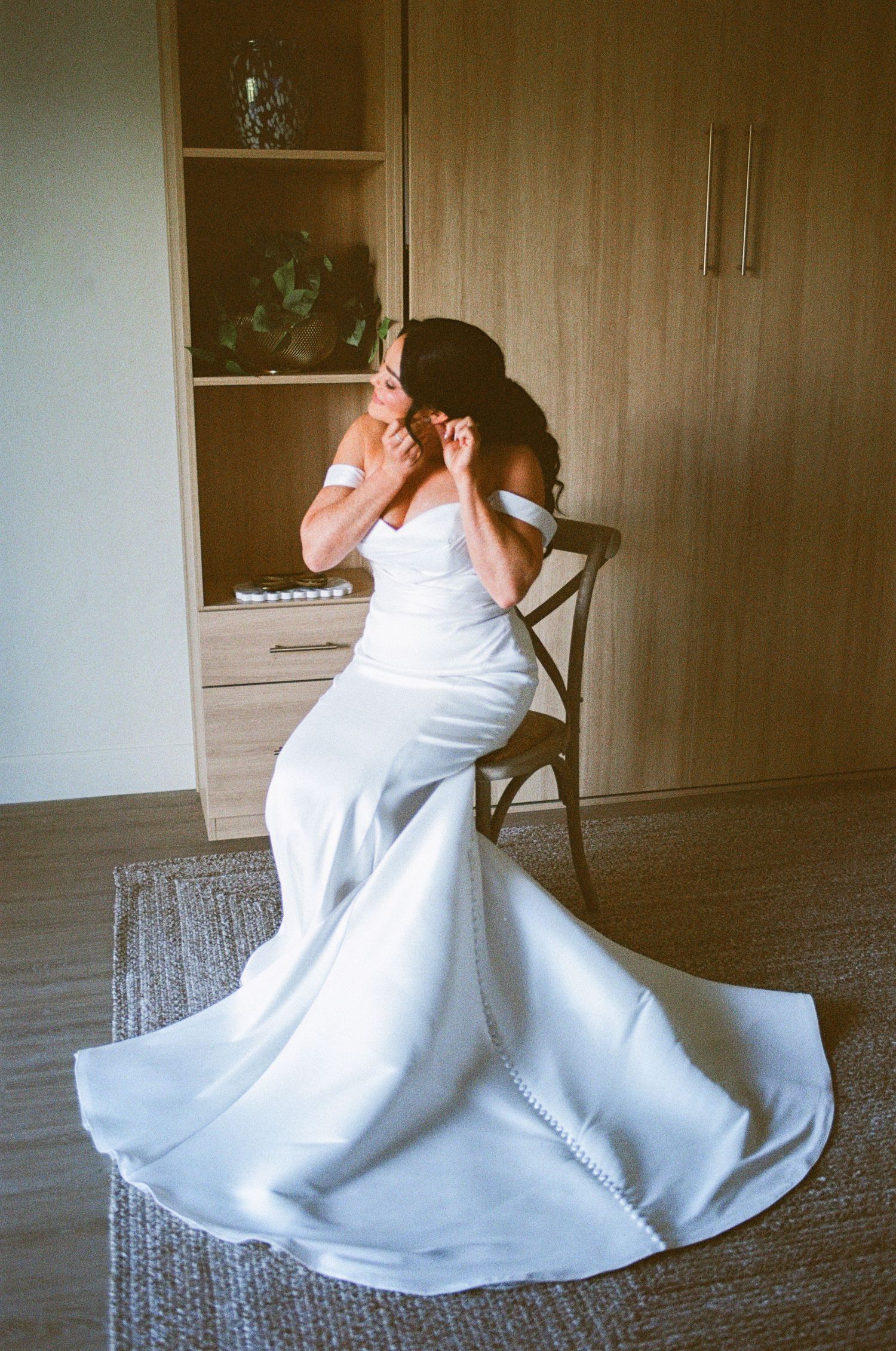 Elegant bride in strapless satin gown adjusting earring while seated in chair.