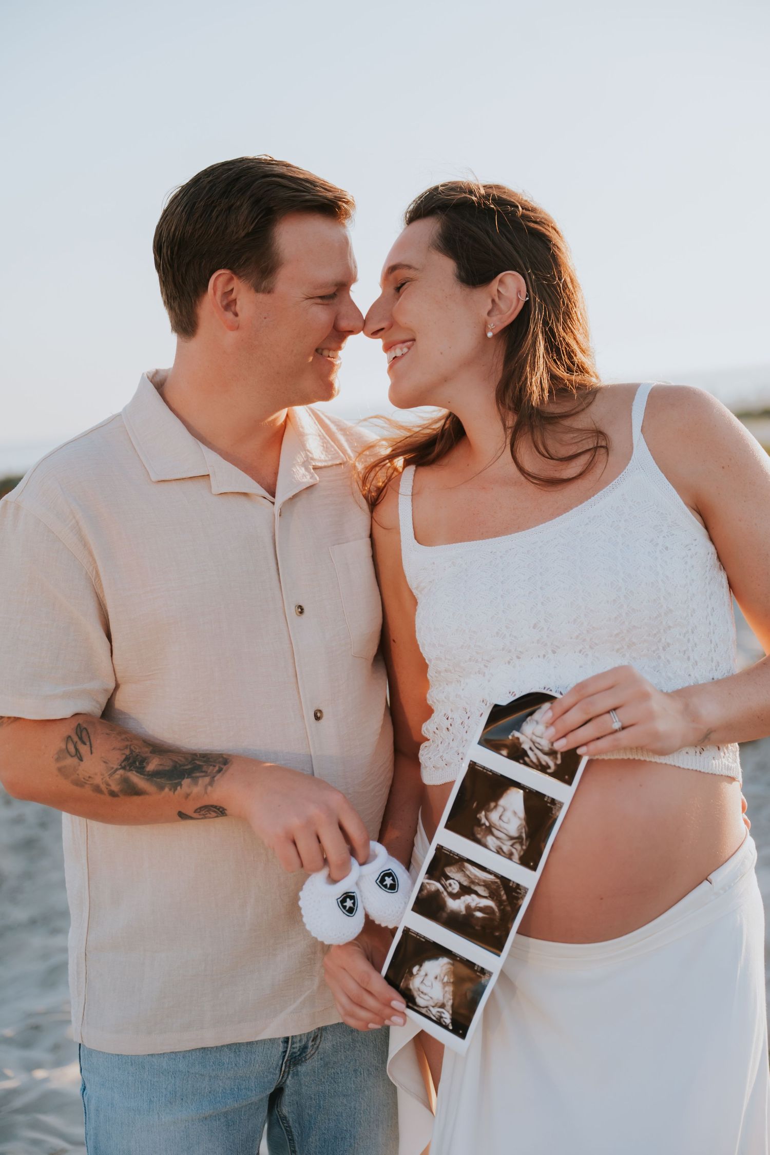 Pregnant couple smiling at each other on beach, holding ultrasound photos and baby booties at golden hour.