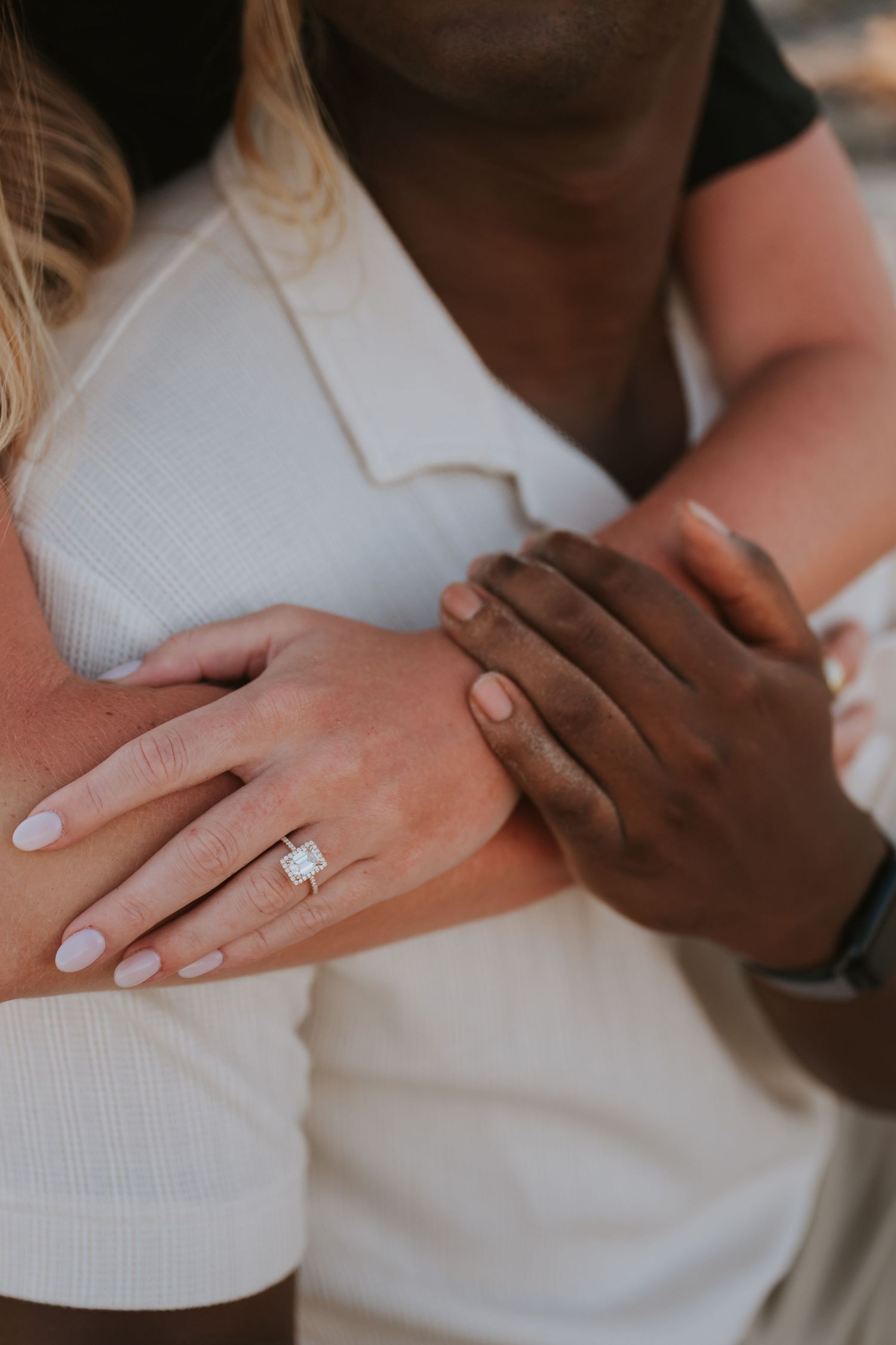 Close-up of an interracial couple embracing, showcasing a sparkling diamond engagement ring on the woman's finger.
