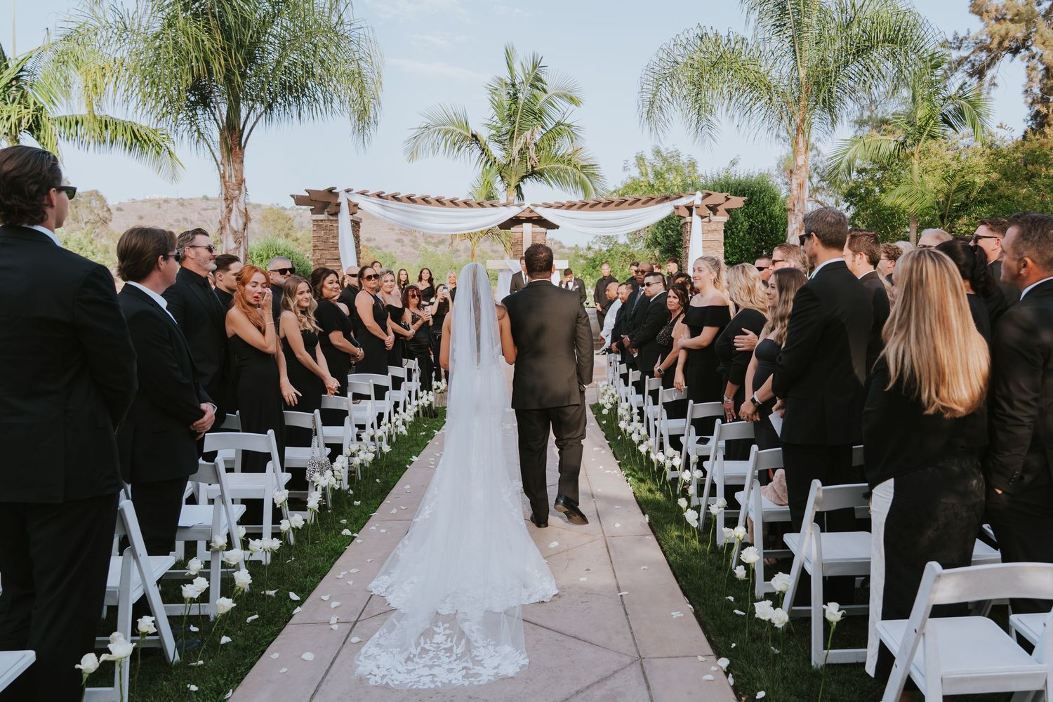 Bride walks down outdoor wedding aisle with palm trees, white chairs, and guests in formal attire at a sunny ceremony.