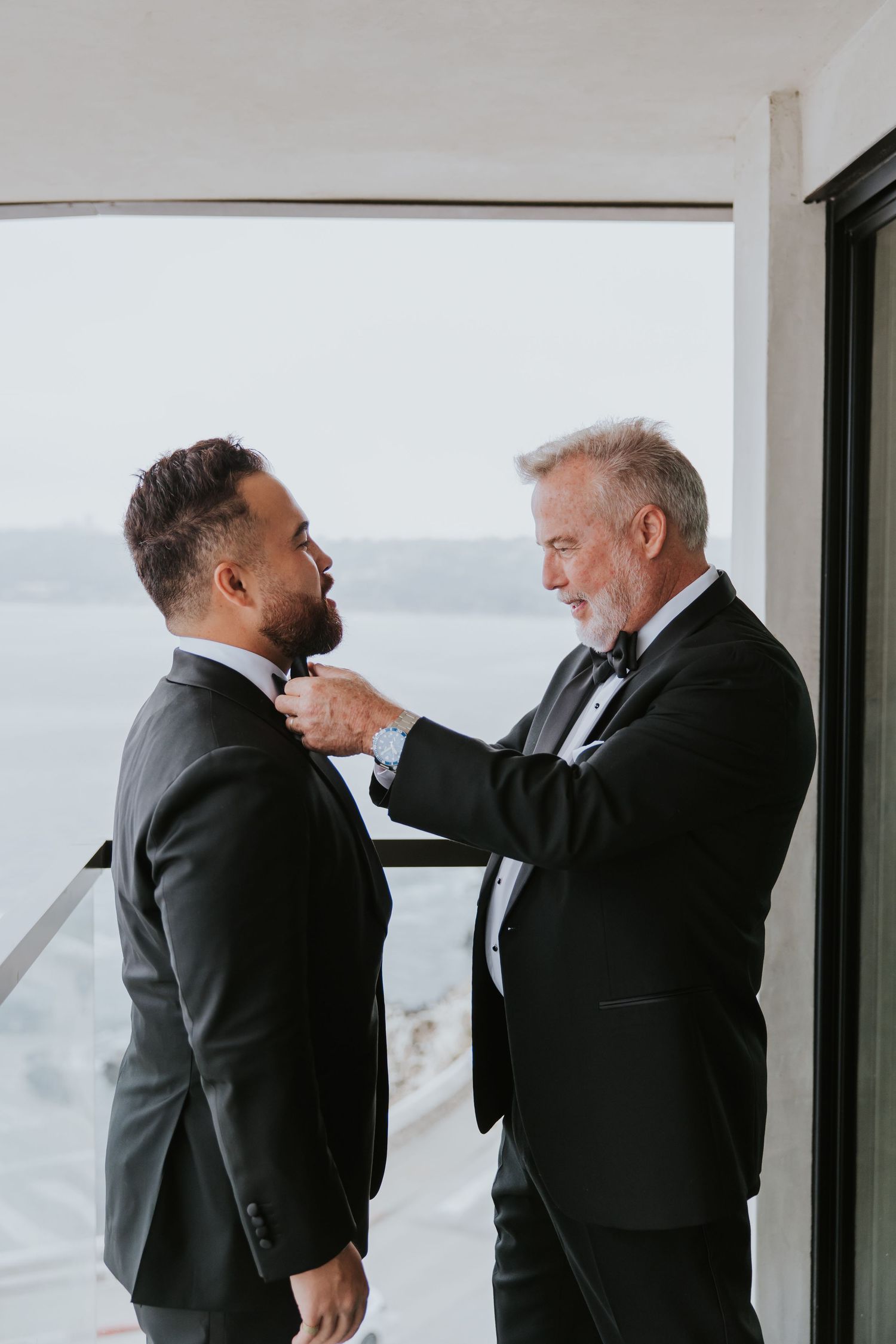 Dad and groom in formal attire adjusting each other's bow ties by a window.