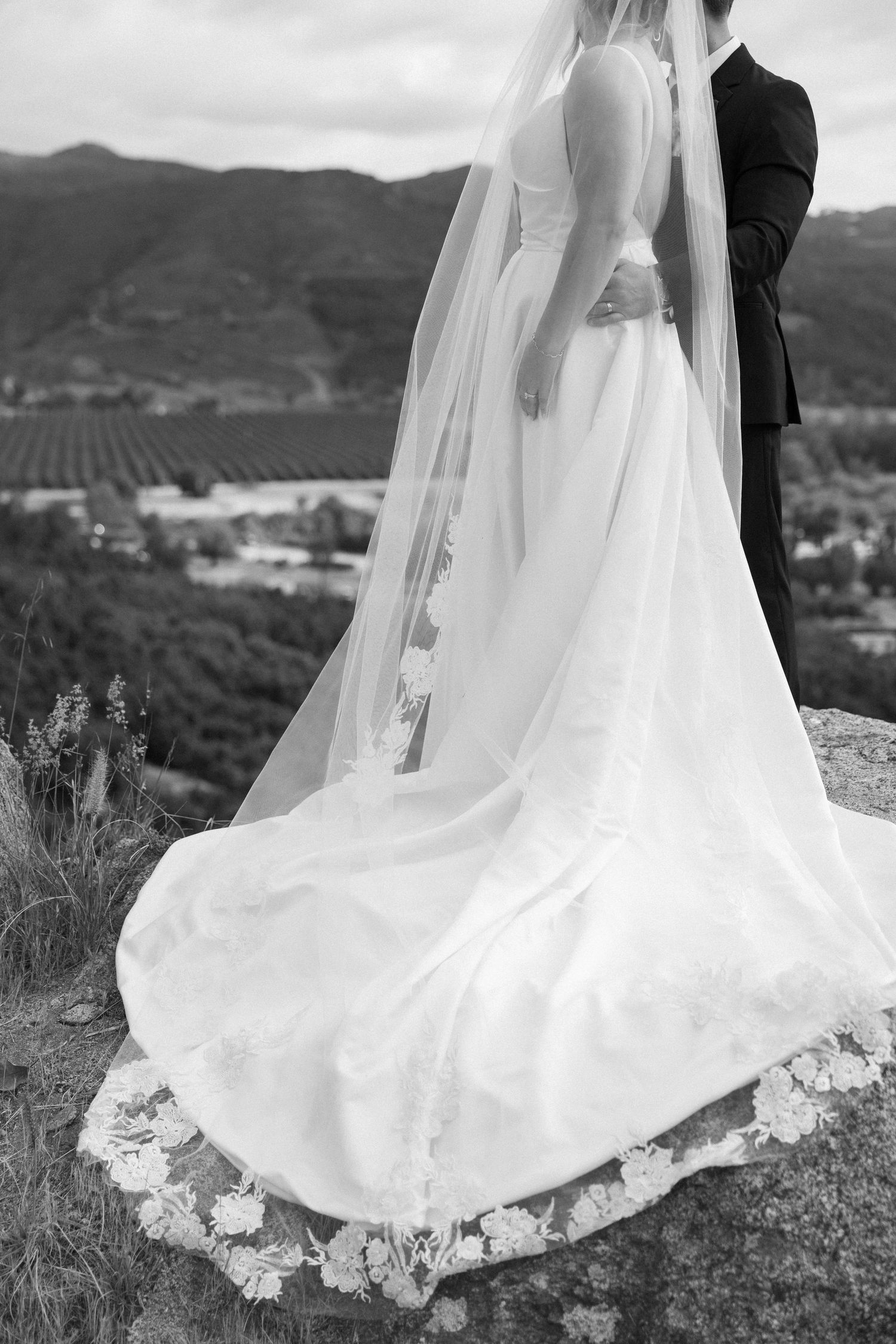 Black and white photo of bride in flowing gown with lace veil and groom overlooking scenic mountain valley.