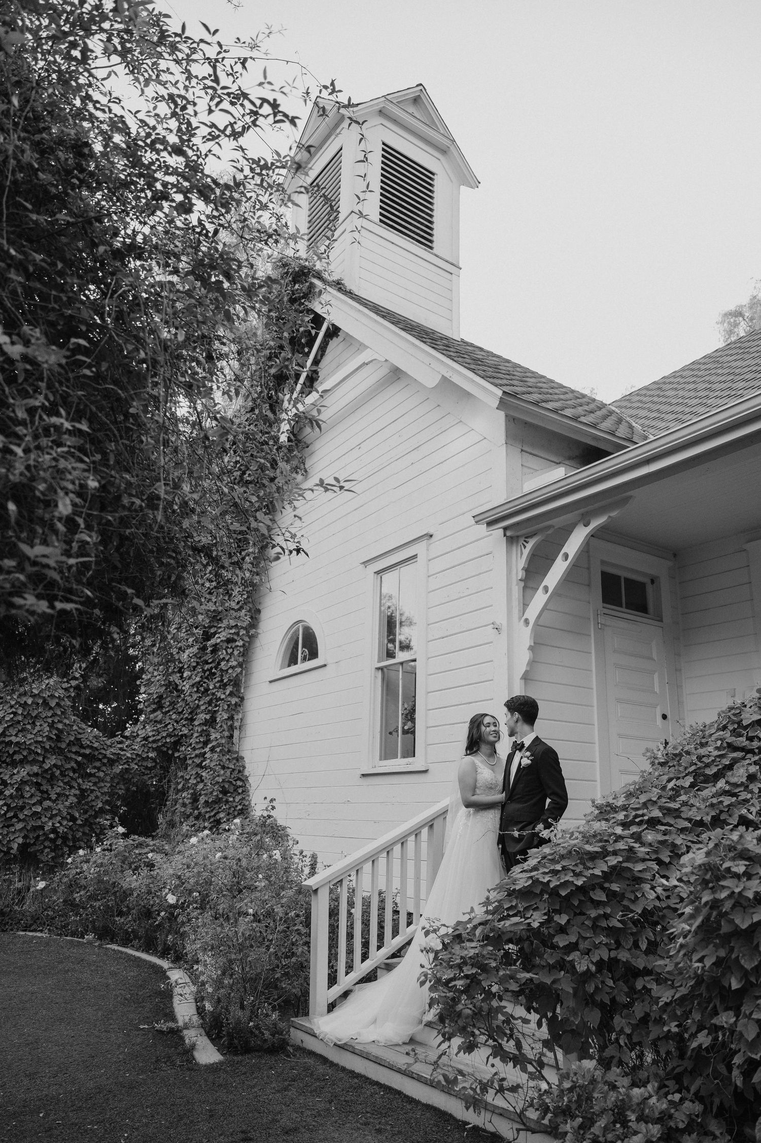 Black and white photo of couple on white chapel steps surrounded by lush greenery.