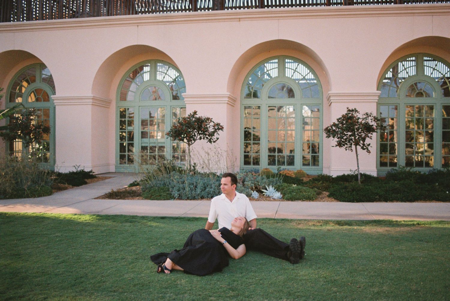 Couple sitting together on green lawn outside elegant arched building with large windows at dusk.