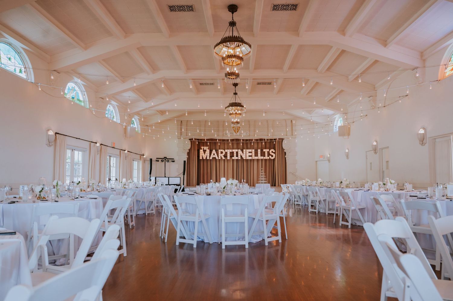 Spacious white wedding reception hall with chandeliers and round tables set for dining.