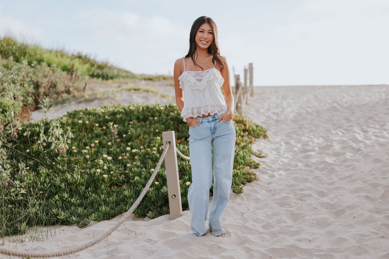 Young woman in white lace top and light blue jeans posing on a sandy beach path with dunes and greenery.