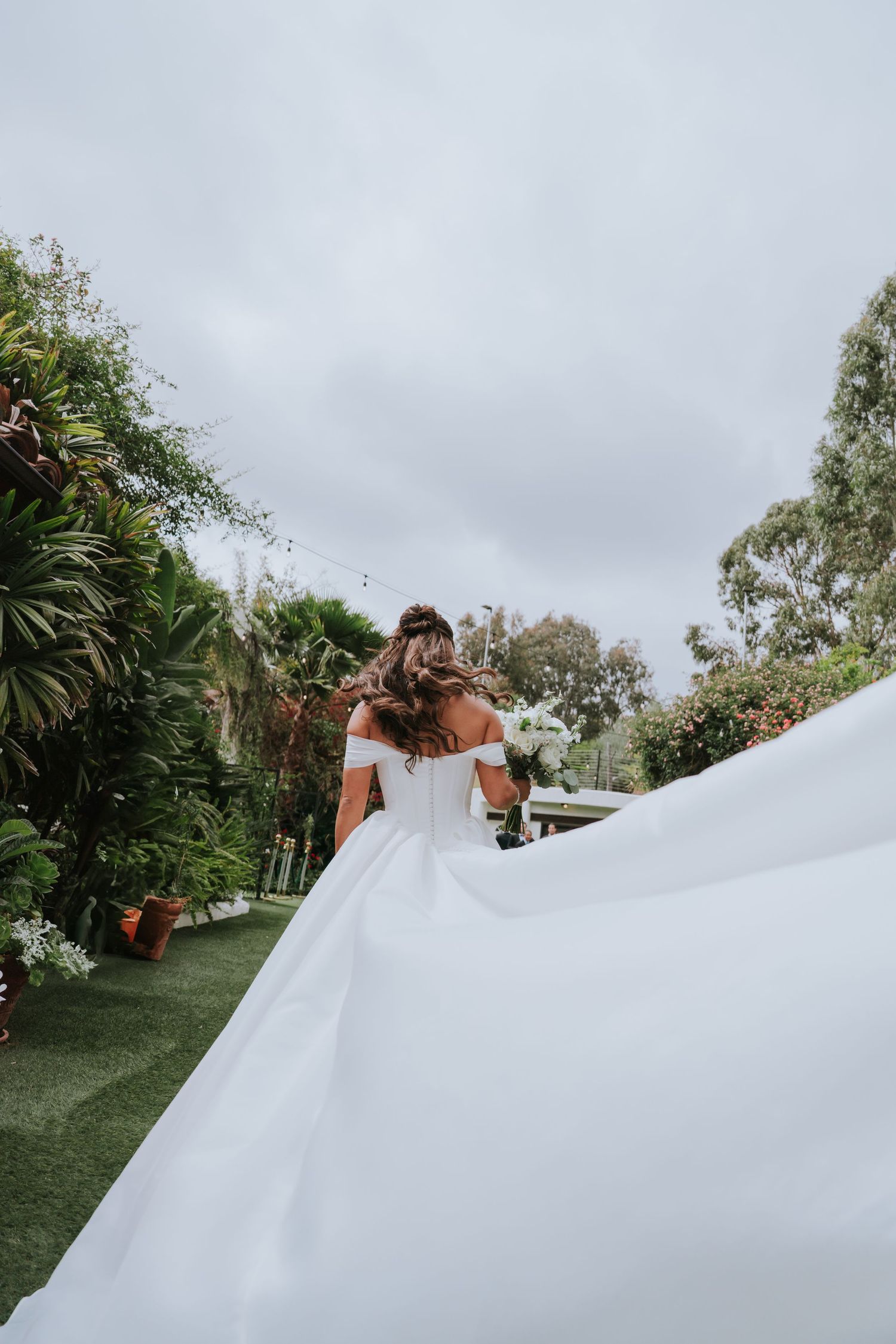 Bride in flowing white off-shoulder gown walks through lush garden, her long train billowing behind her on her wedding day.
