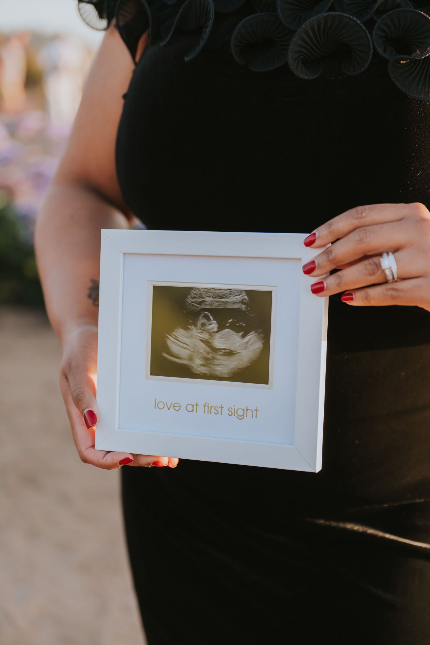 Pregnant woman holding framed ultrasound photo with love at first sight text, wearing black dress with red nails.