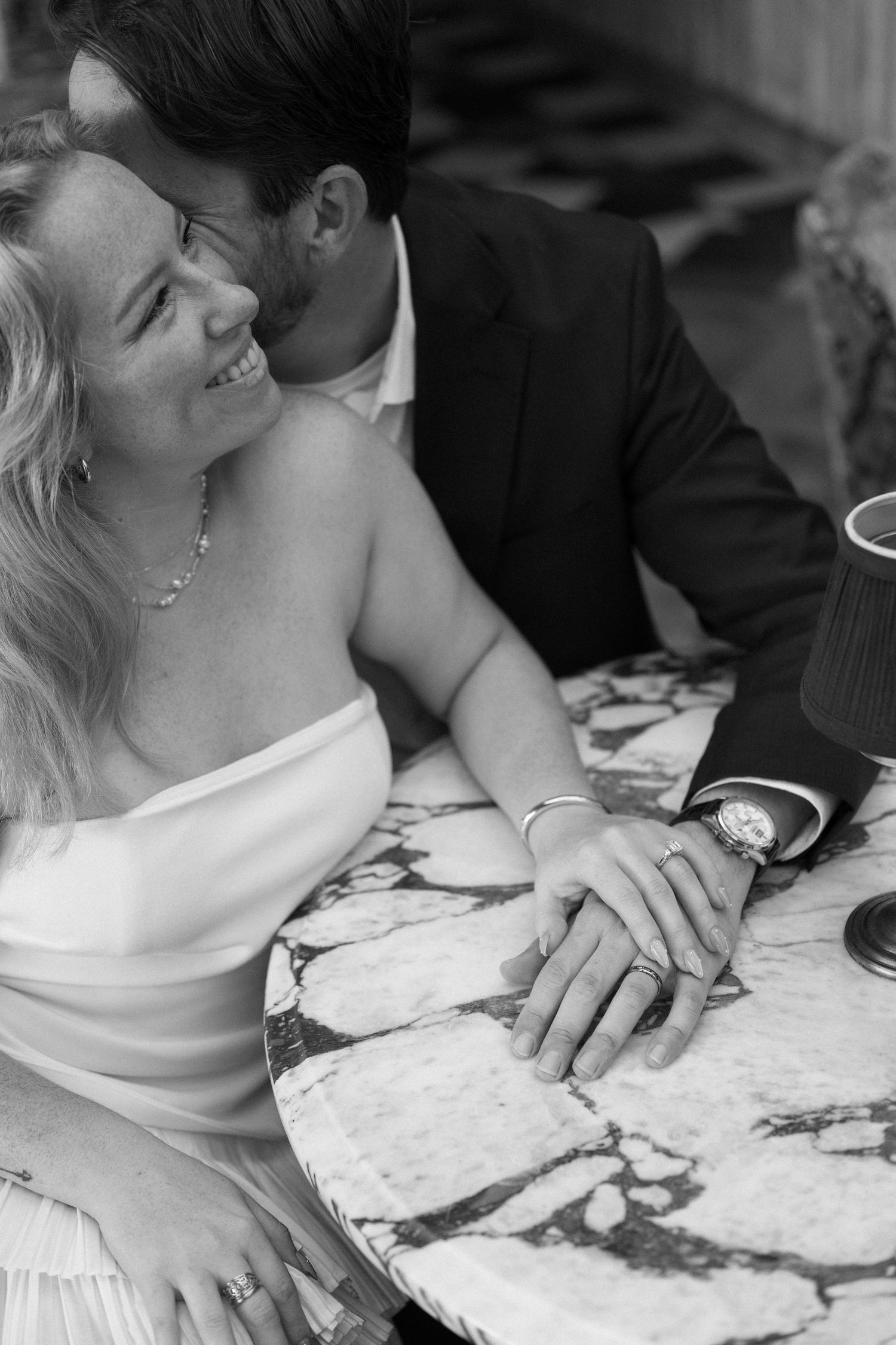 Black and white photo of a smiling bride and groom holding hands at a marble table.