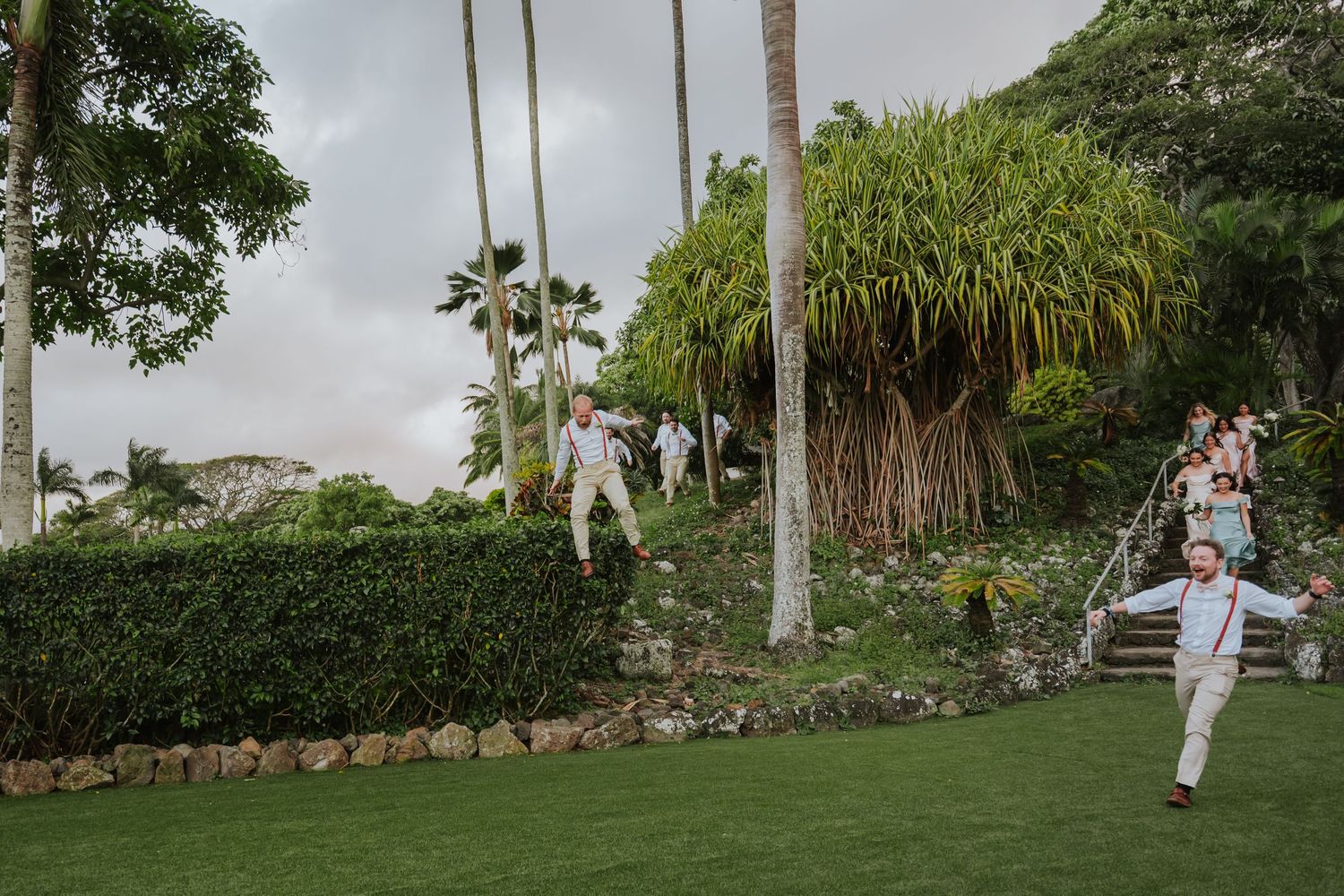 Wedding guests run playfully down stone steps in tropical garden setting.
