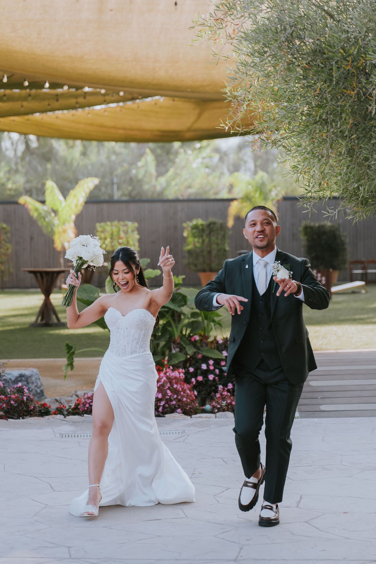 Wedding couple performs fun dance moves on outdoor patio with string lights and greenery in background.