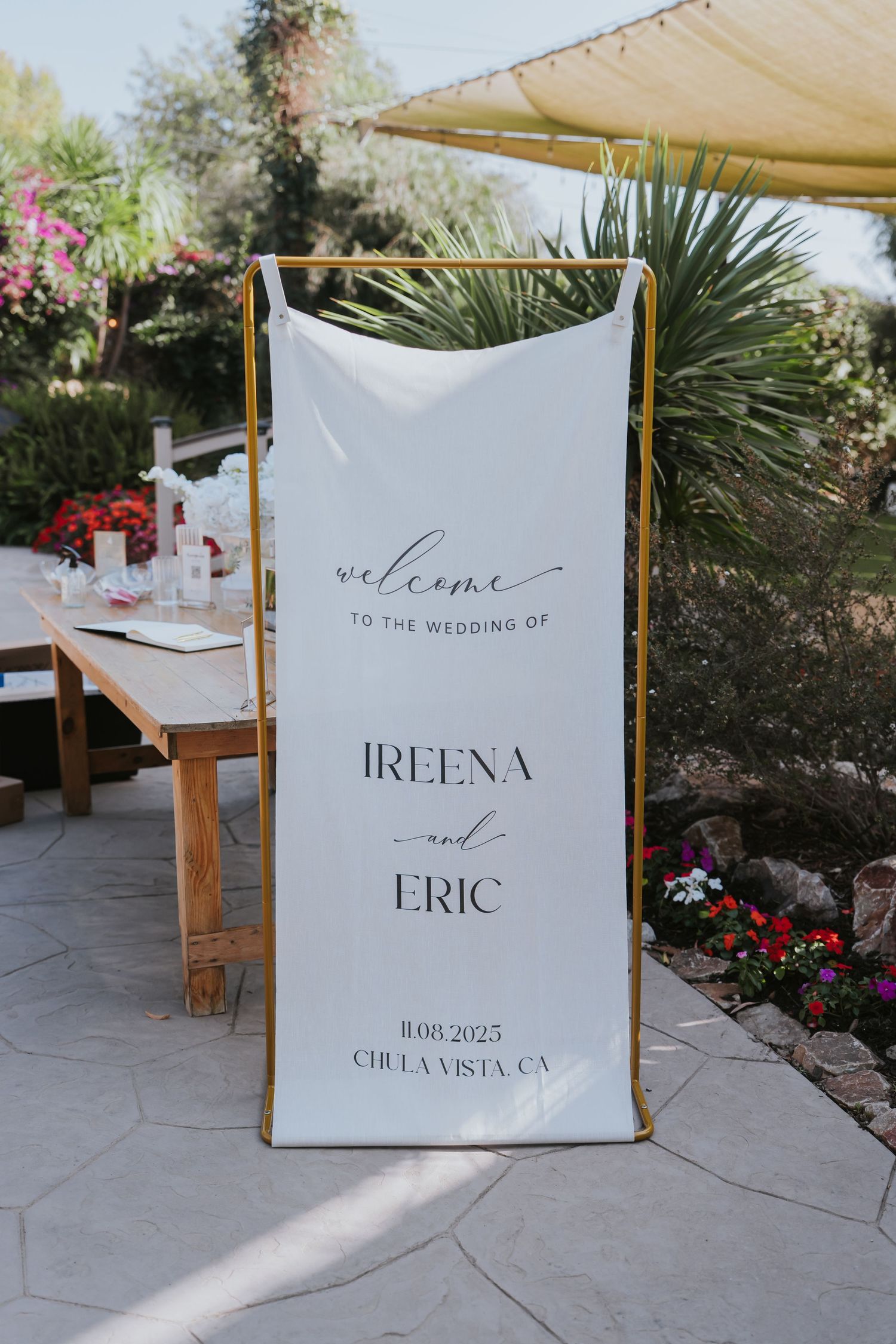 Elegant white wedding welcome sign with gold frame displays couple's names and date at an outdoor garden venue.