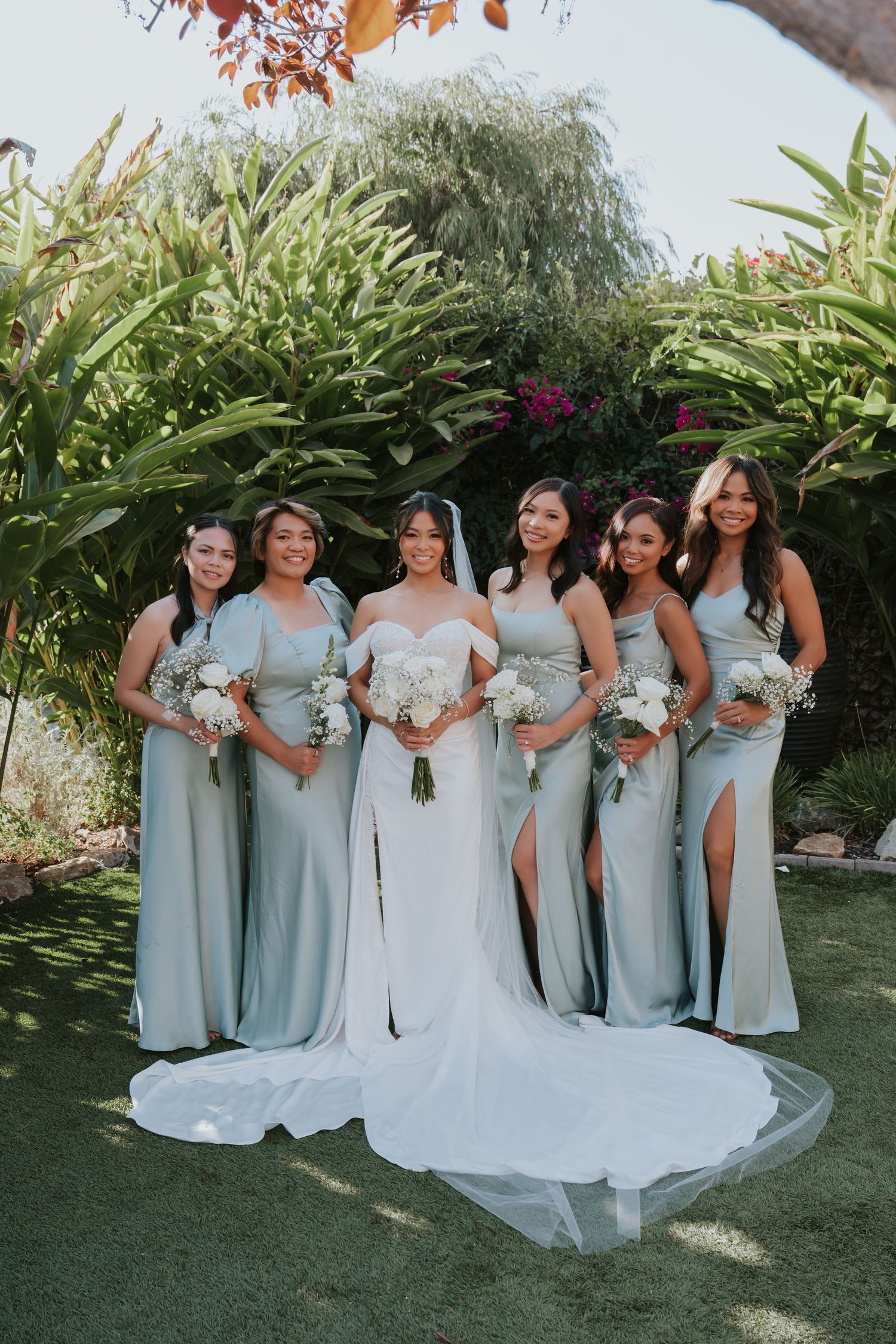 Bridal party in matching pale blue bridesmaid dresses with thigh slits pose with bride in white gown in garden setting.