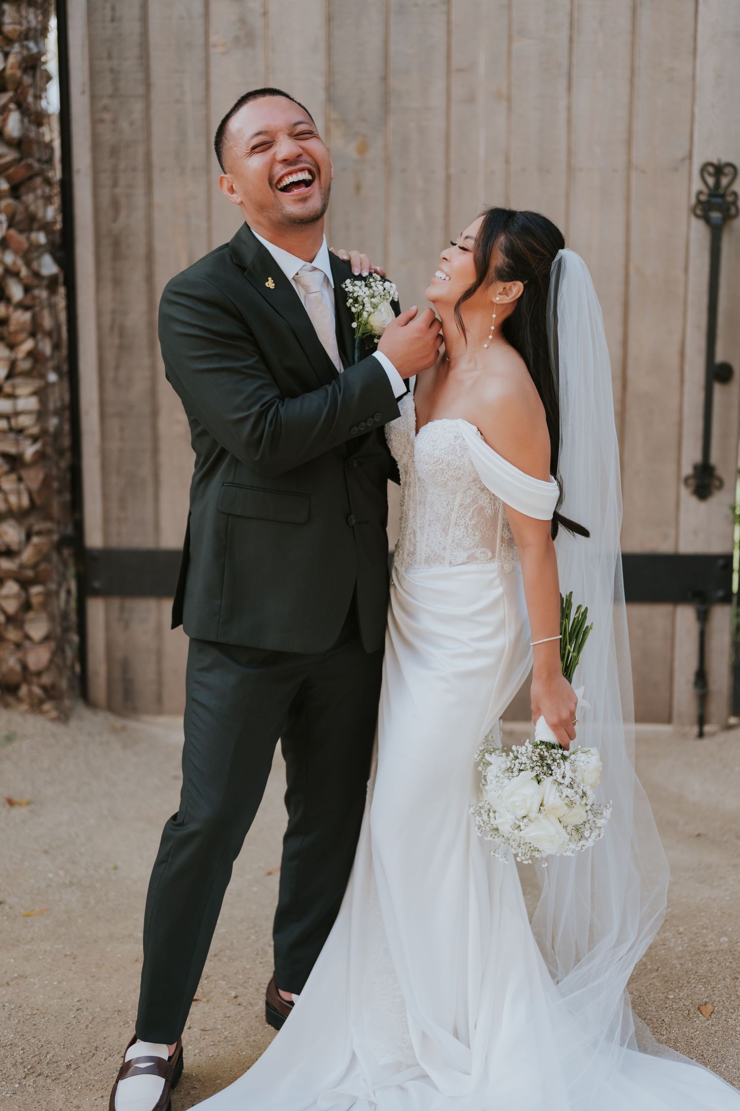 A joyful wedding couple shares a candid moment against a stone wall backdrop.