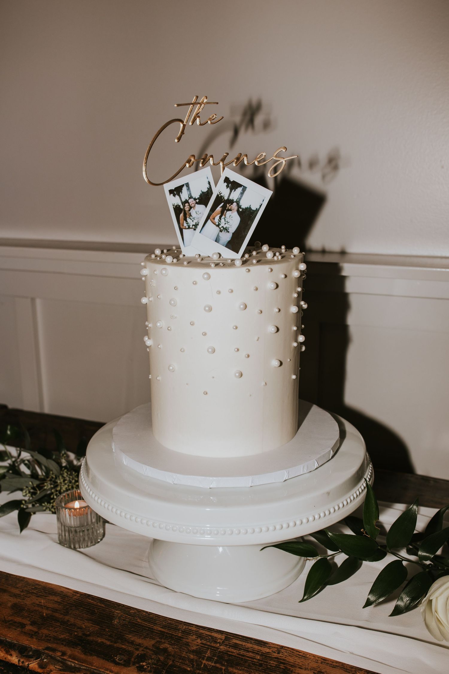 A white wedding cake decorated with pearl dots and topped with photos sits on an elegant cake stand with greenery.