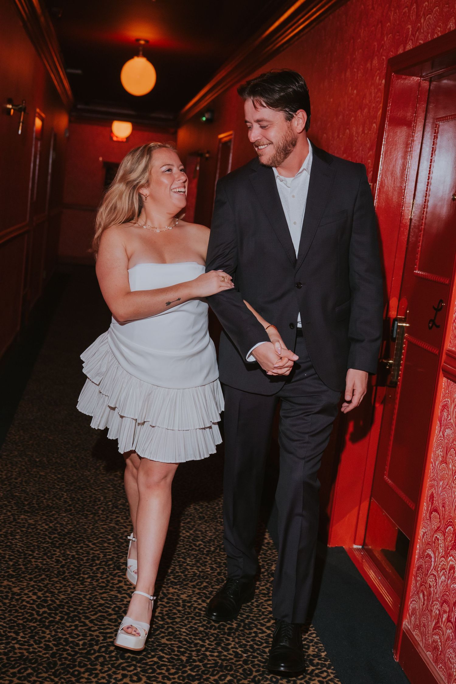 Bride in white ruffled mini dress holds groom's arm as they walk through a red-walled hotel corridor.