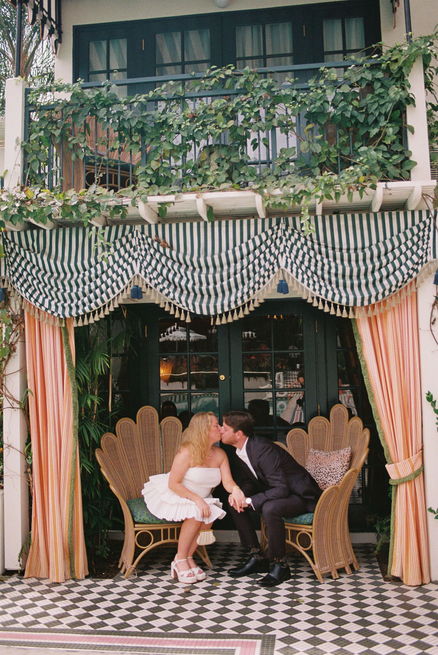 Newlyweds share a romantic kiss seated in wicker chairs under a striped canopy at elegant venue.