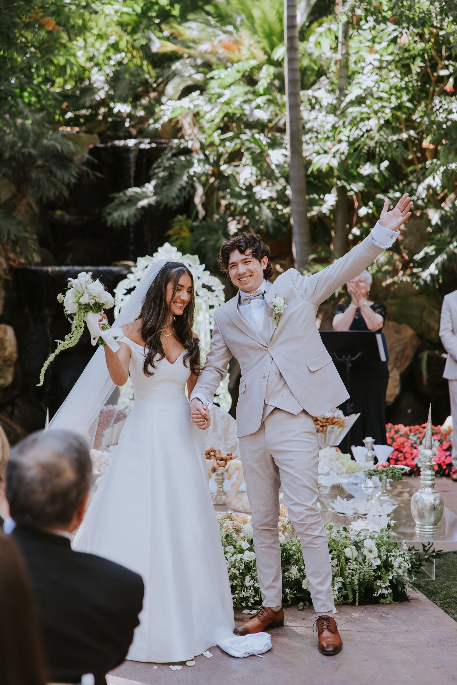 A joyful wedding celebration in a garden setting with people dancing and celebrating in white and beige attire.
