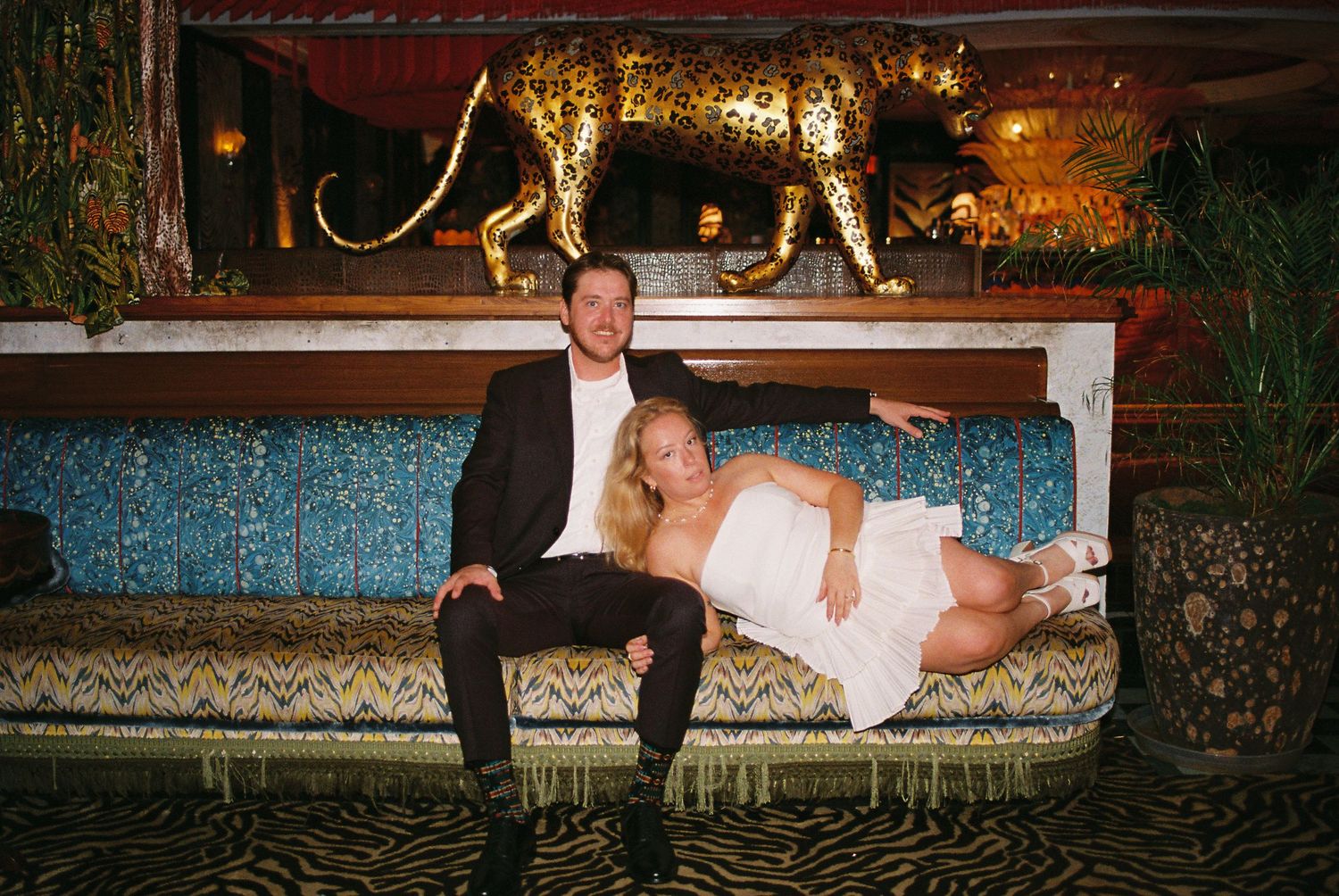 Newlyweds posing on ornate sofa beneath gold leopard statue in stylish venue.