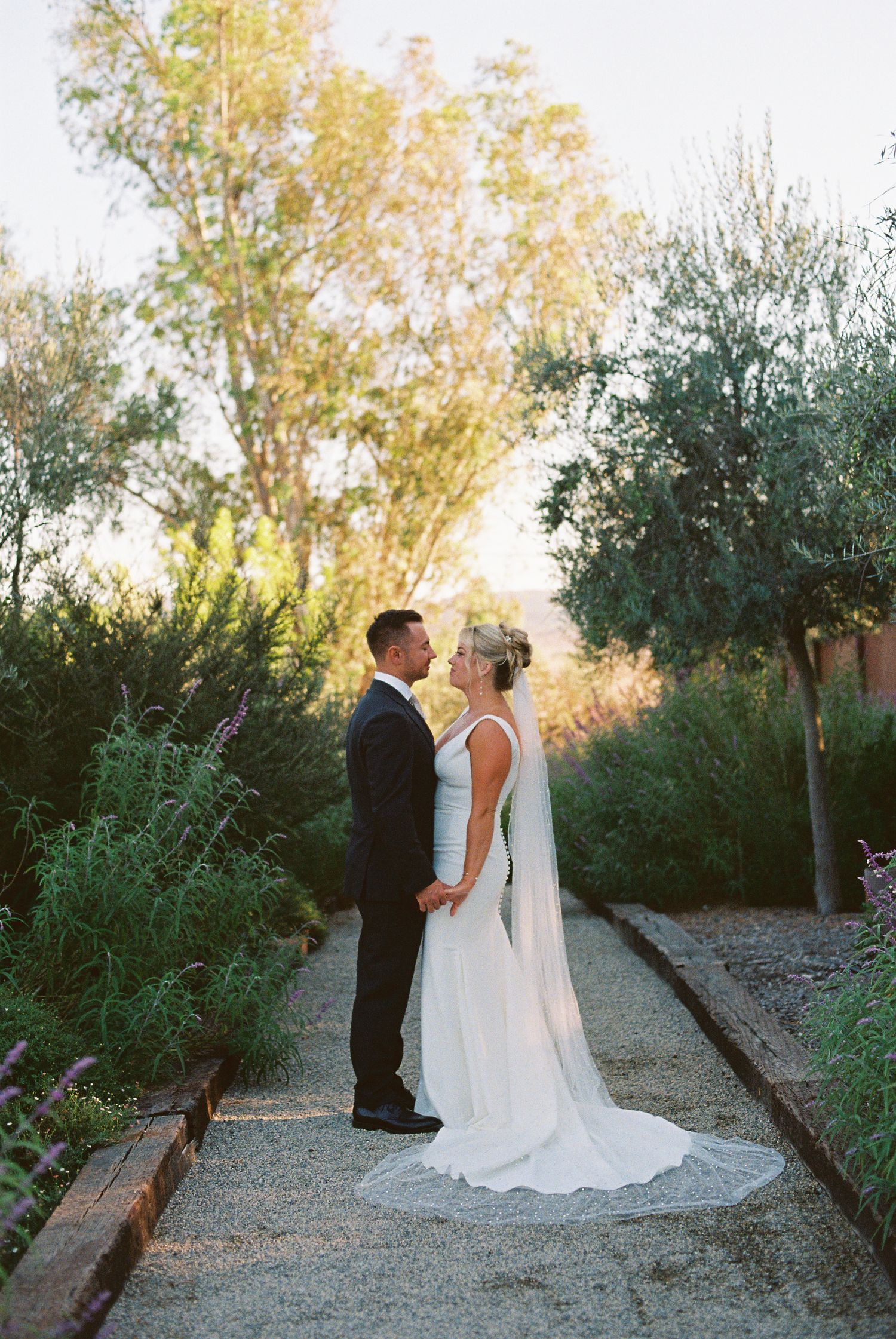 Bride and groom share a romantic moment on a garden path at golden hour, surrounded by lush greenery.