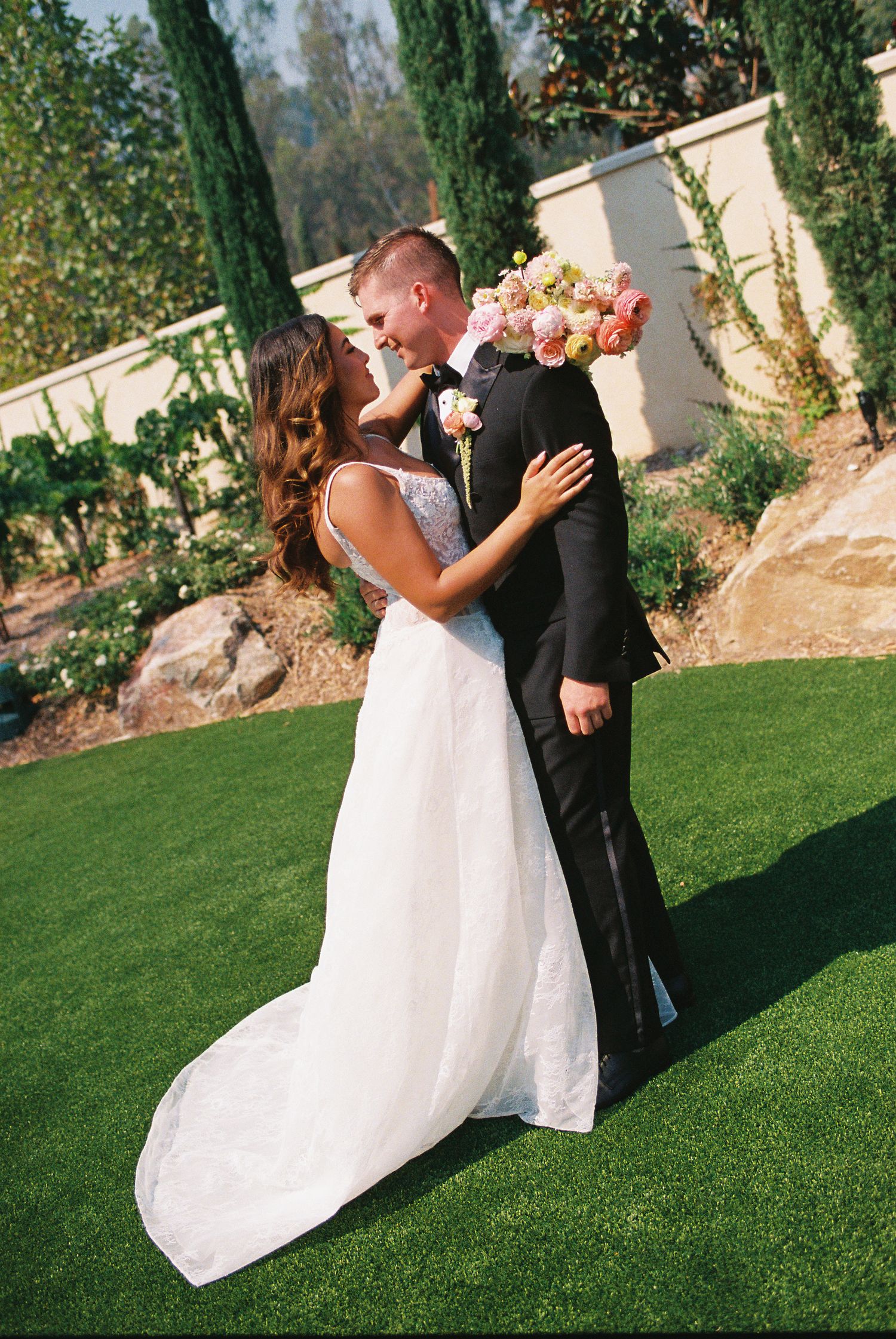 Bride in white gown and groom in black tuxedo share a romantic embrace outdoors on green lawn with pink bouquet.