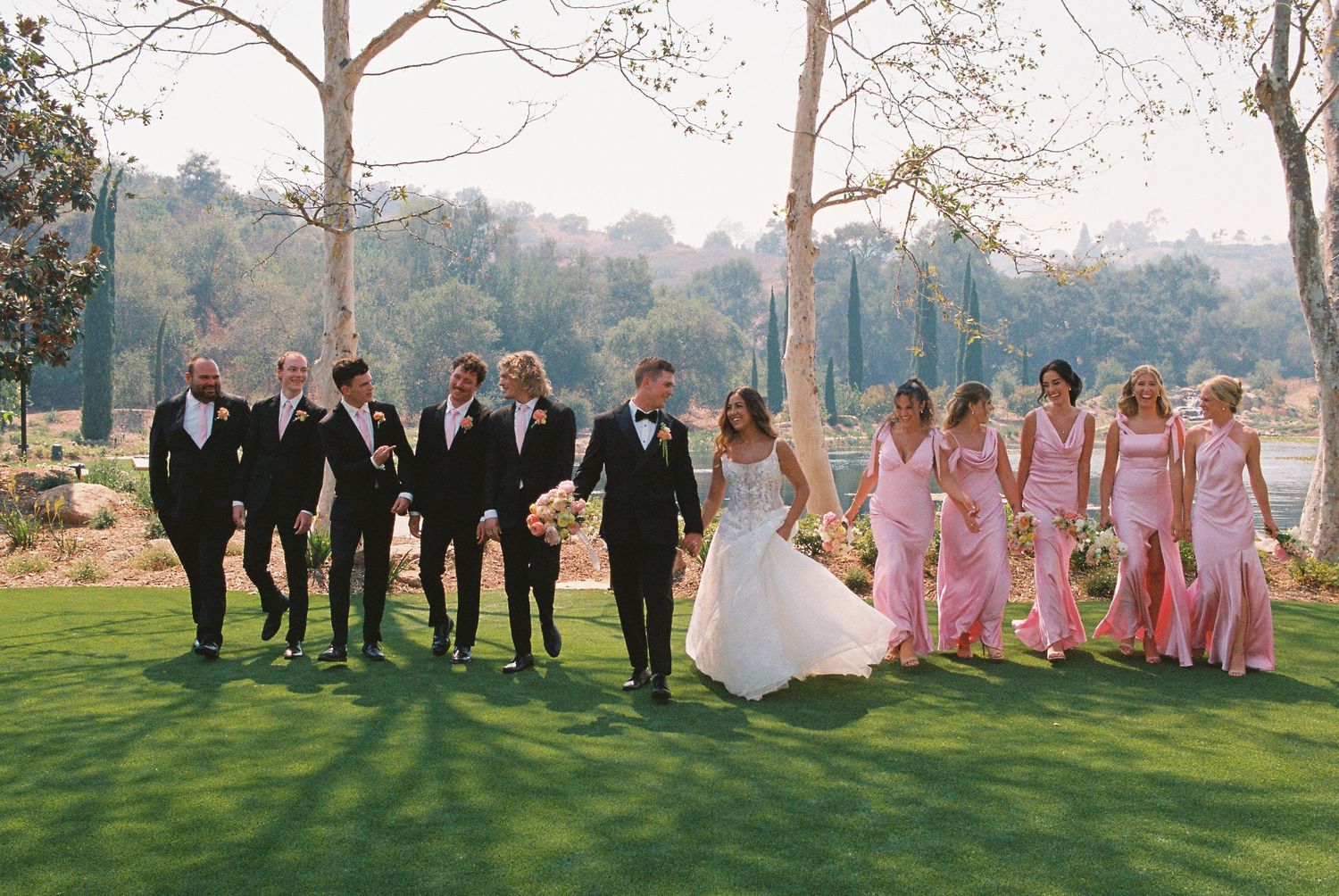Wedding party walks on green lawn, groomsmen in black tuxedos and bridesmaids in pink gowns with trees behind.
