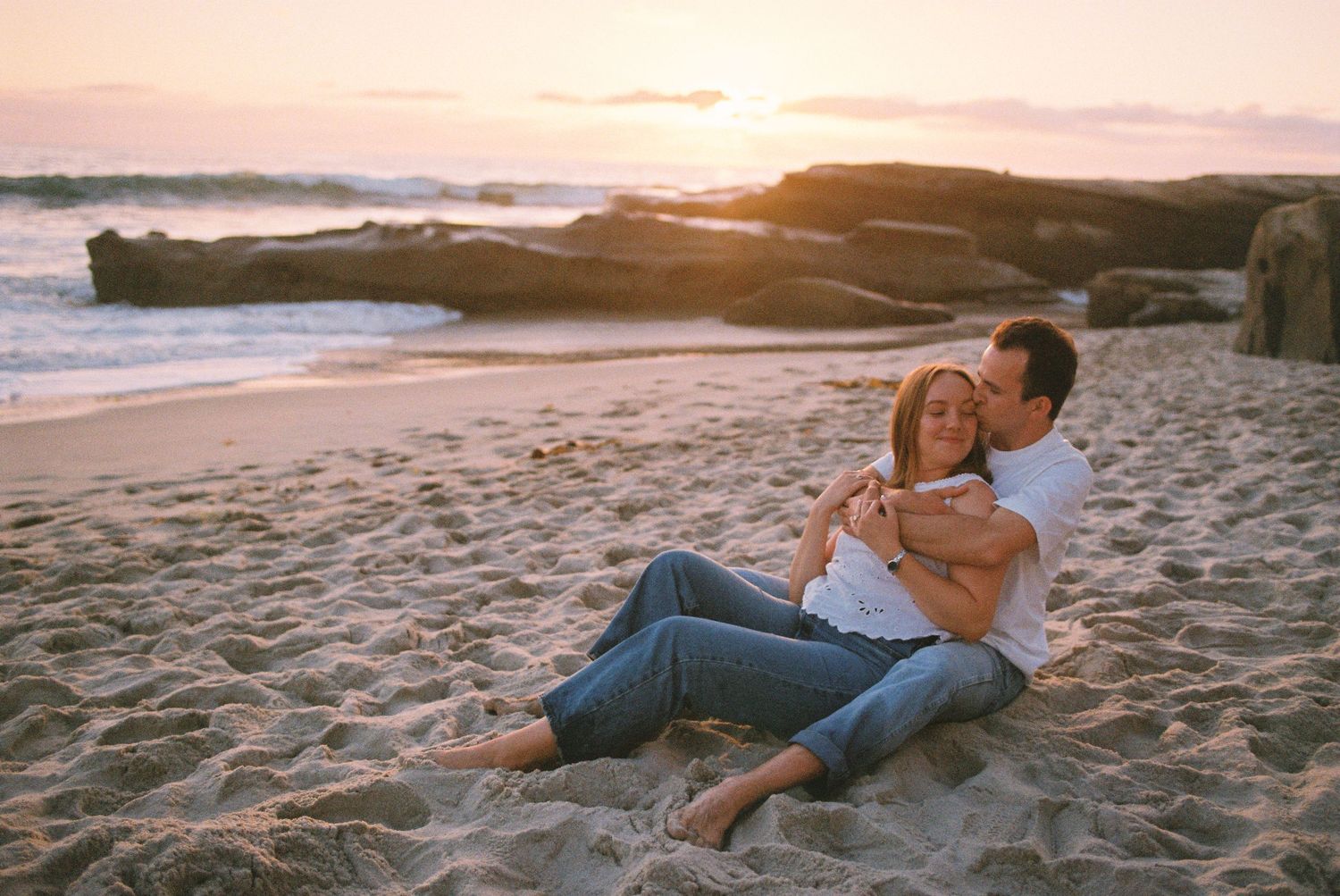 Couple sitting together on sandy beach at sunset, embracing romantically with ocean waves and rocks in background.
