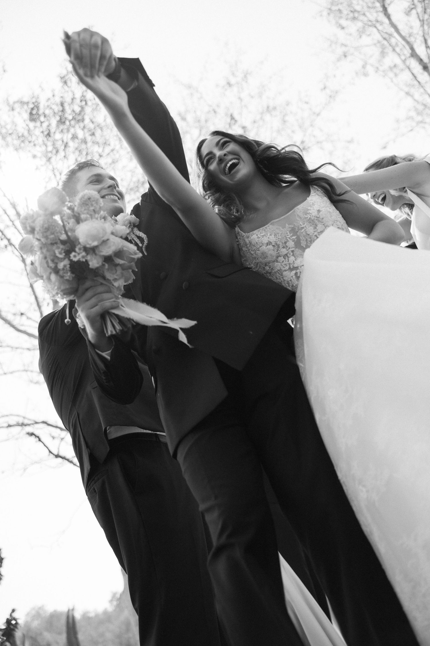 Black and white photo of joyful newlyweds celebrating, bride in gown holding bouquet, groom in suit, arms raised outdoors.