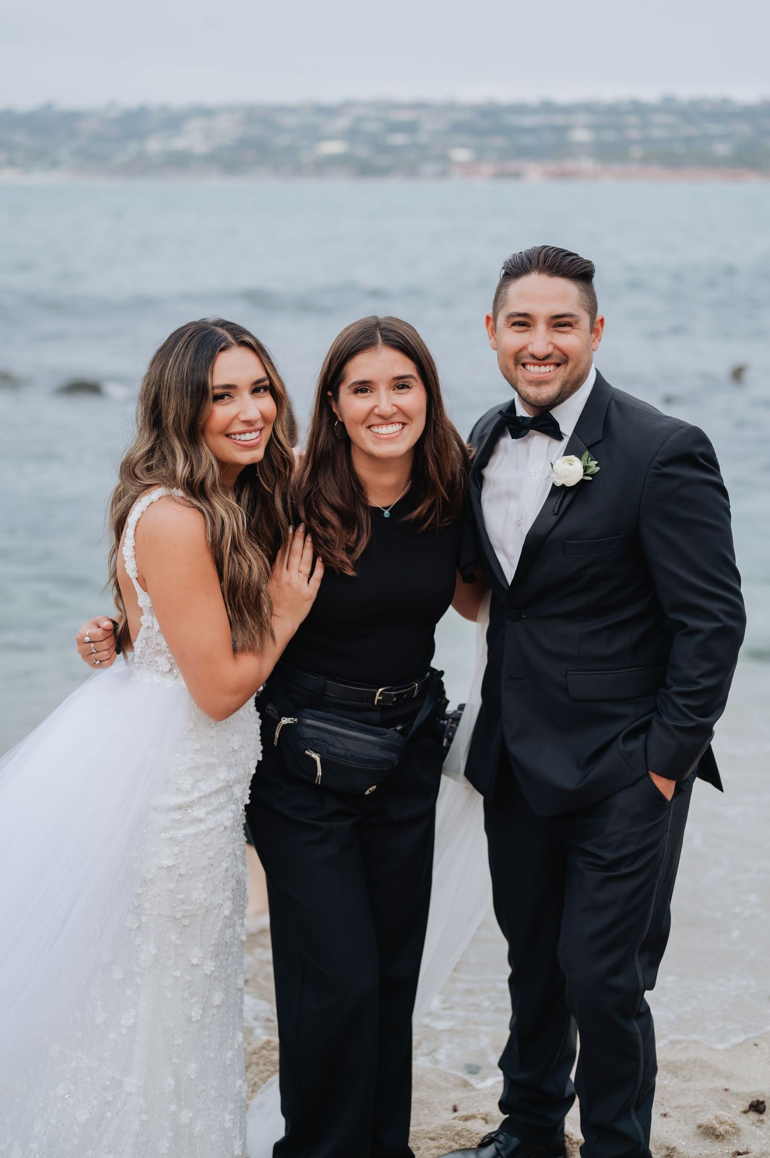 Bride in white gown and groom in black tuxedo pose with wedding coordinator on a scenic beachside location.
