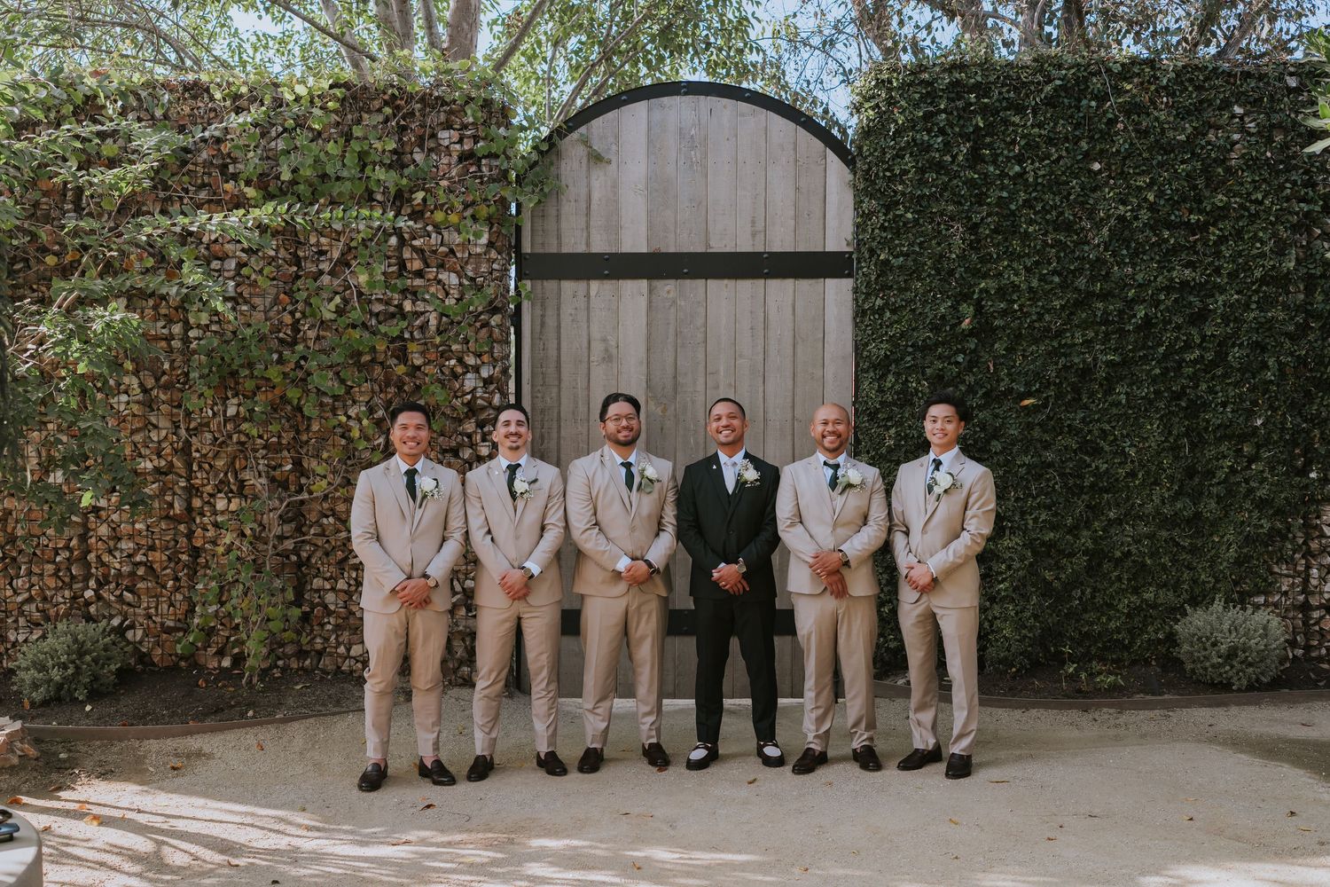 Wedding party in tan suits and black suit standing in front of rustic wooden gate with ivy-covered wall.