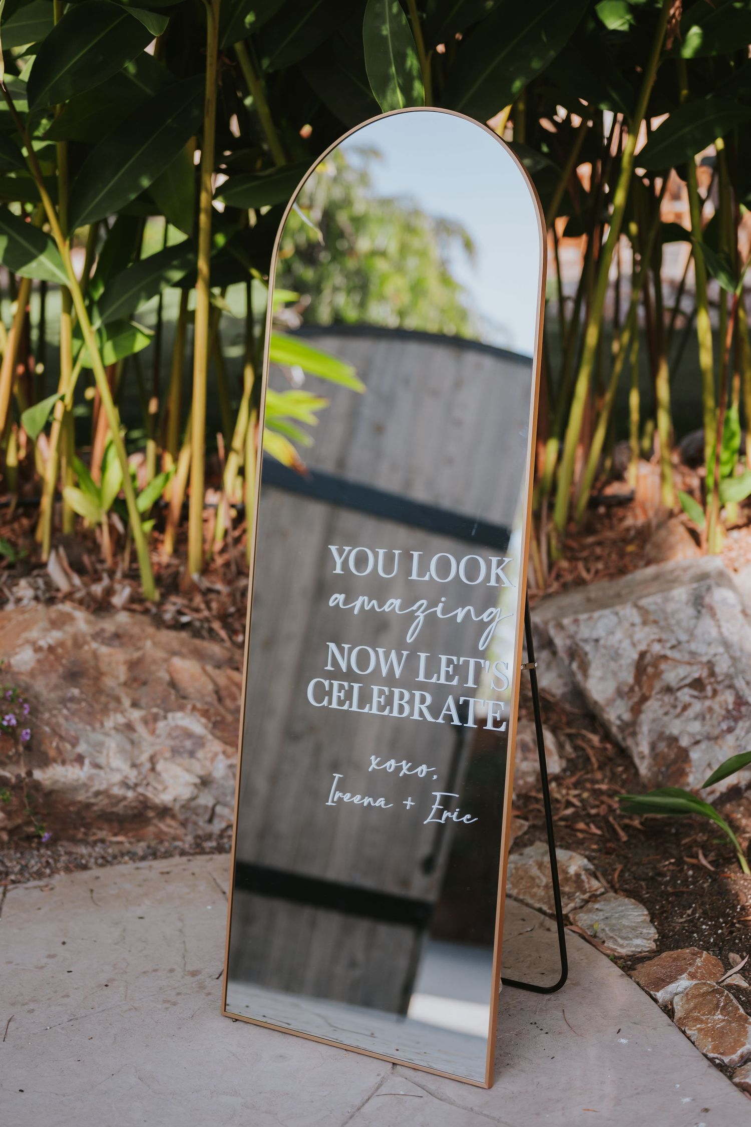 A decorative mirror with text reading 'You look amazing now let's celebrate' displayed in an outdoor garden setting.