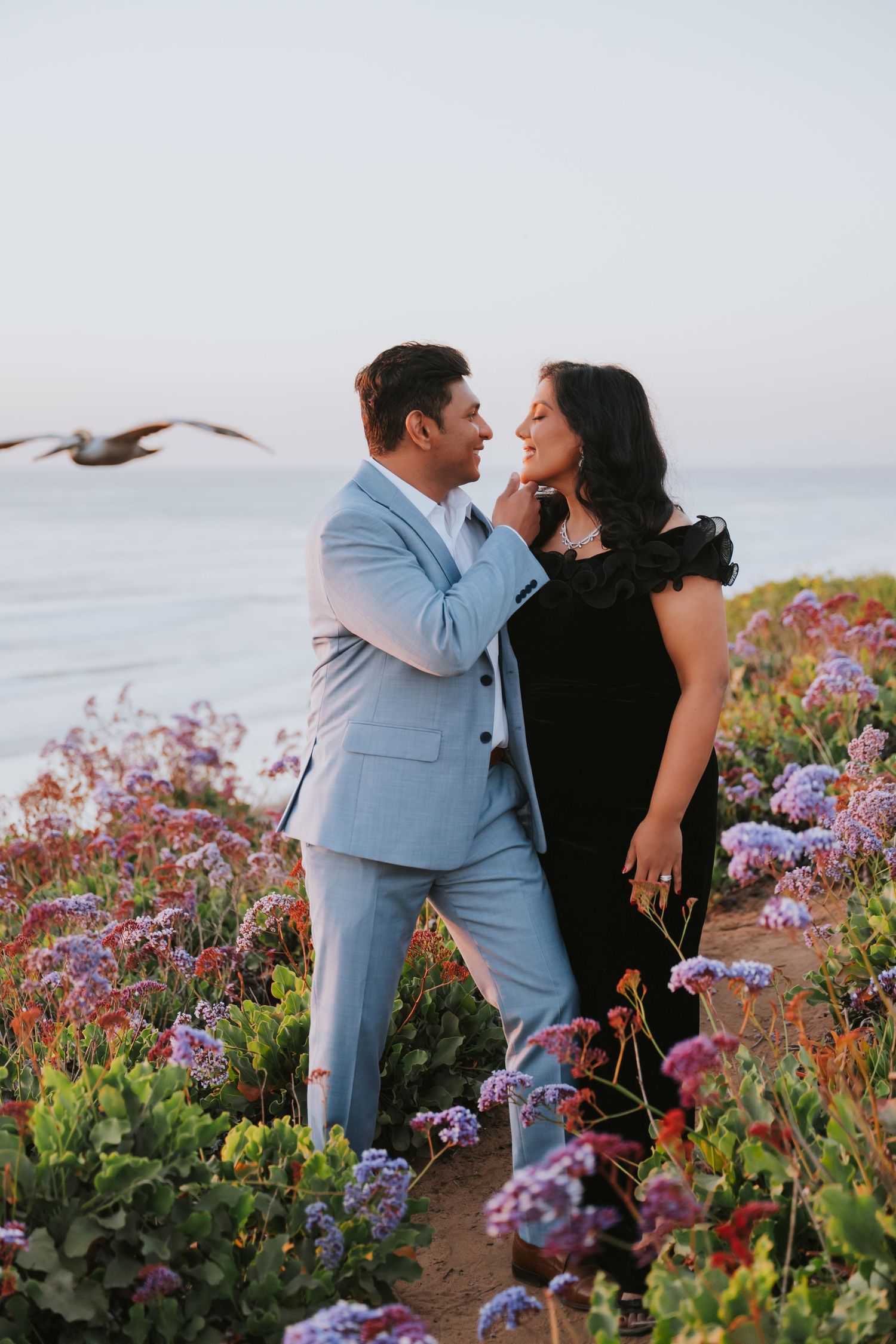 Couple embracing among purple wildflowers on a coastal cliffside at sunset, man in light blue suit, woman in black dress.