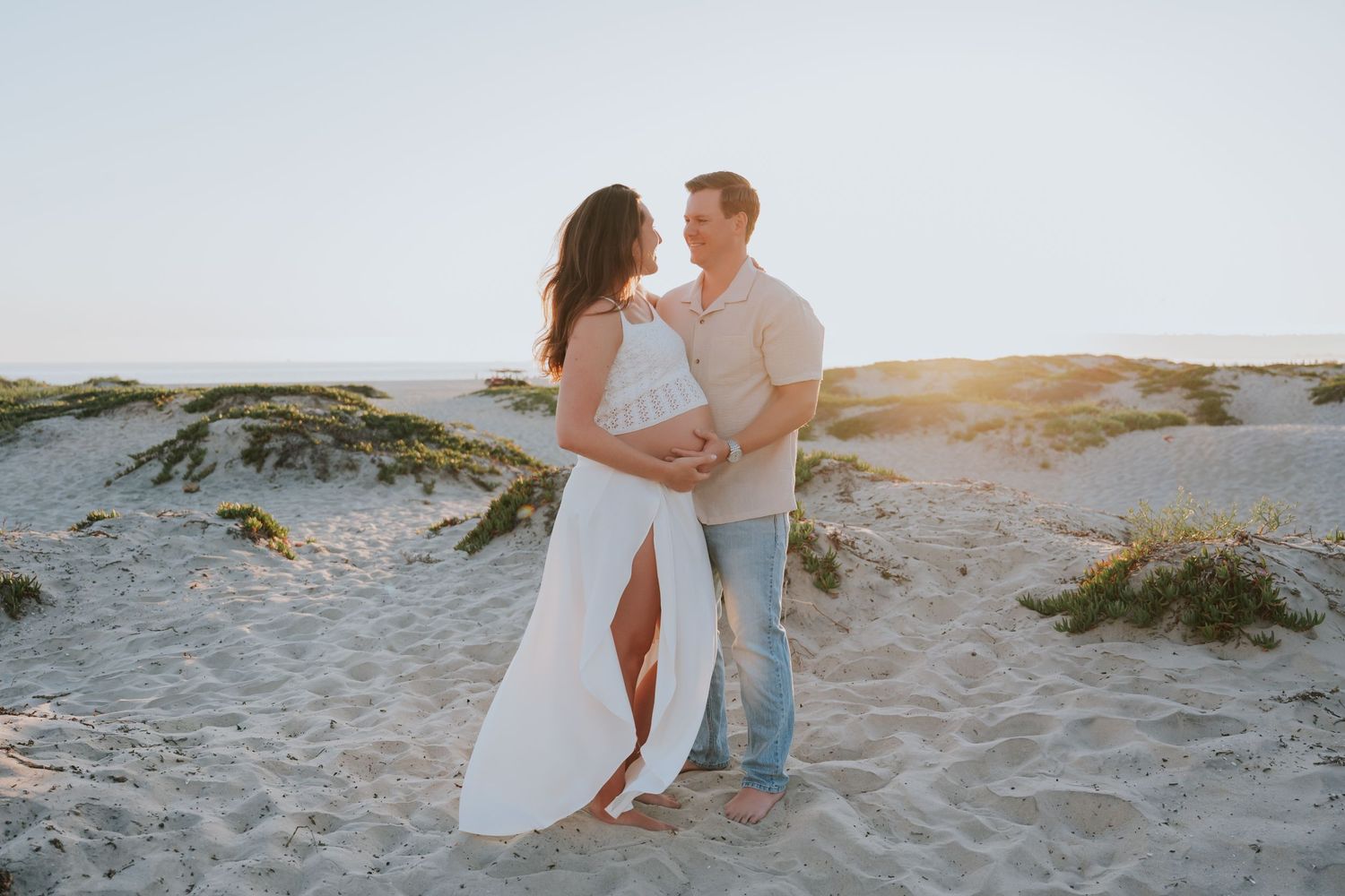 Couple embracing on sandy beach dunes at golden hour, woman in white dress, man in casual outfit, soft warm sunlight.