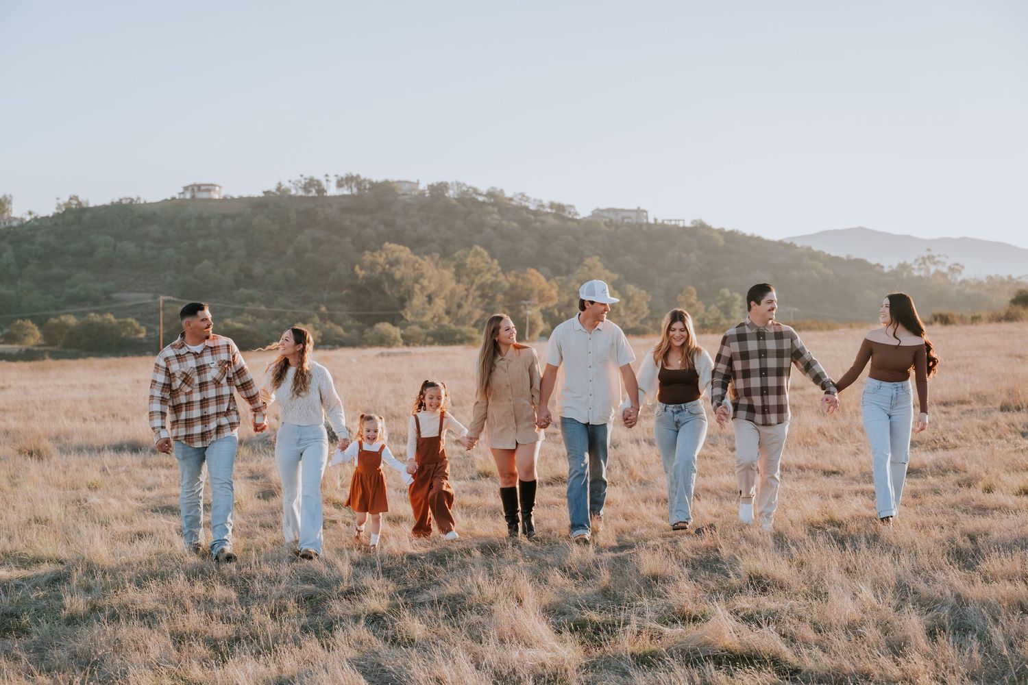 Large family holding hands walking through golden field with rolling hills in background on sunny day.