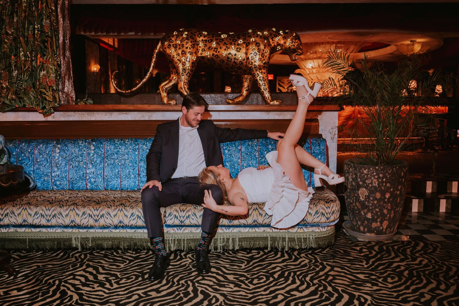 Couple laughing on ornate blue sofa, woman playfully kicking legs up, golden leopard statue backdrop in stylish venue.